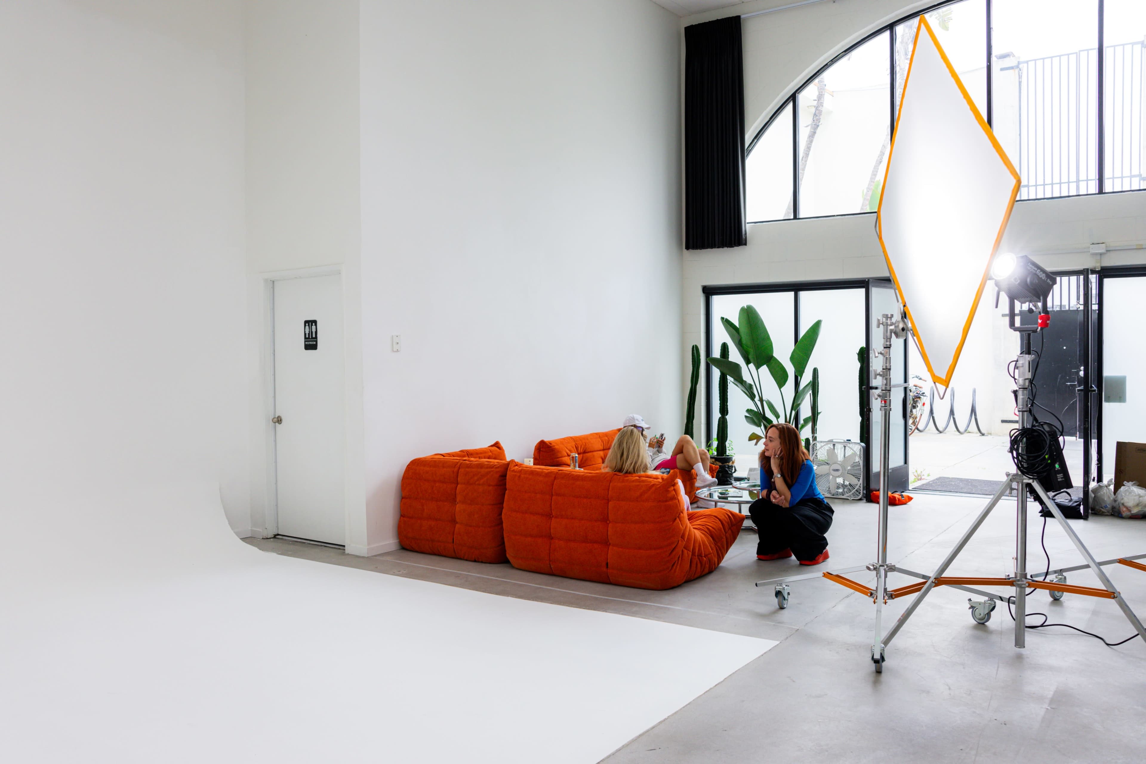 A well-lit studio features an orange sectional sofa, a large white backdrop, and a lighting setup with a reflective umbrella near a glass door.
