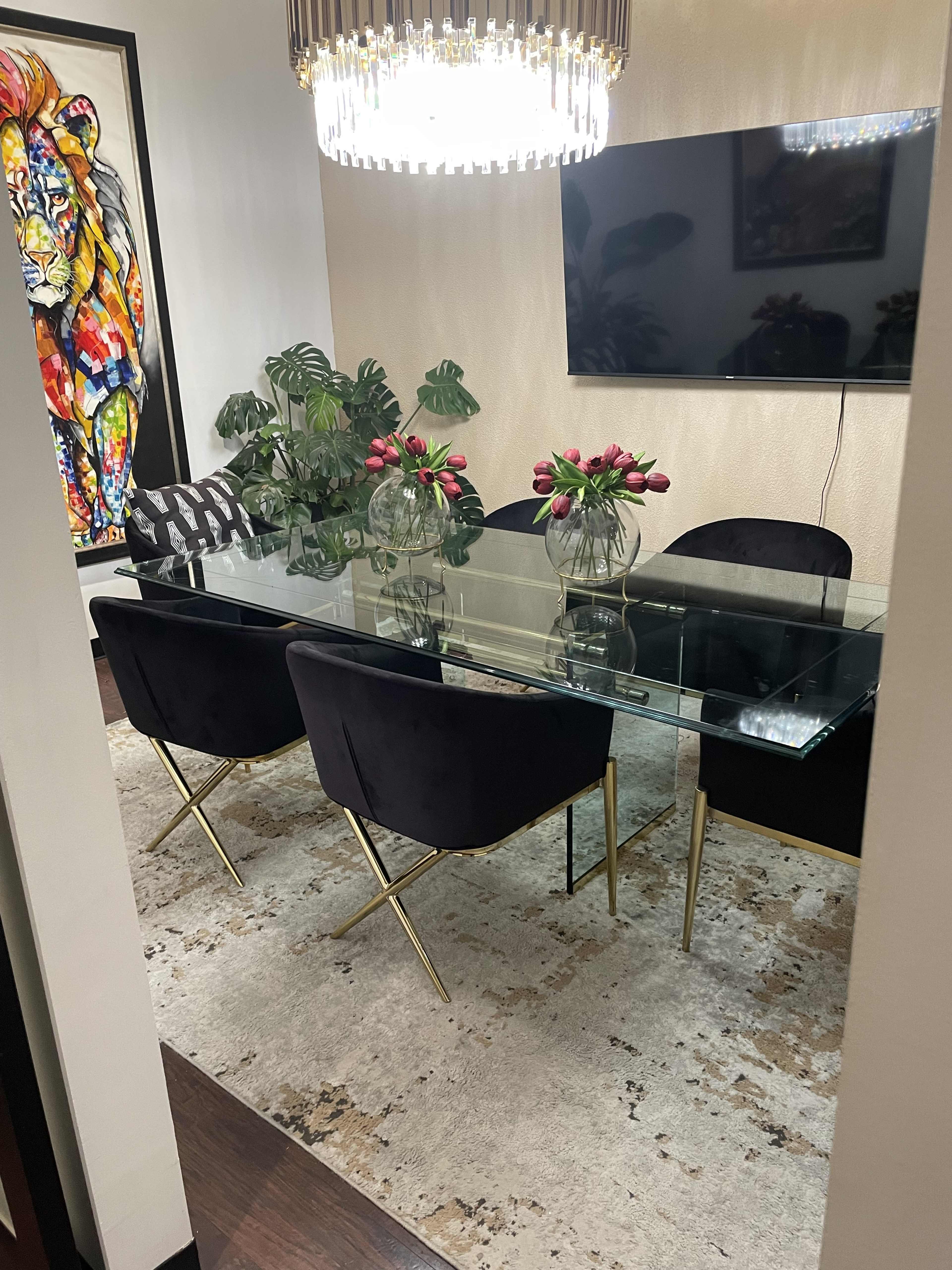 A modern dining room with a glass table, velvet chairs, and decorative plants, framed by a colorful lion painting and a flat-screen TV on the wall.