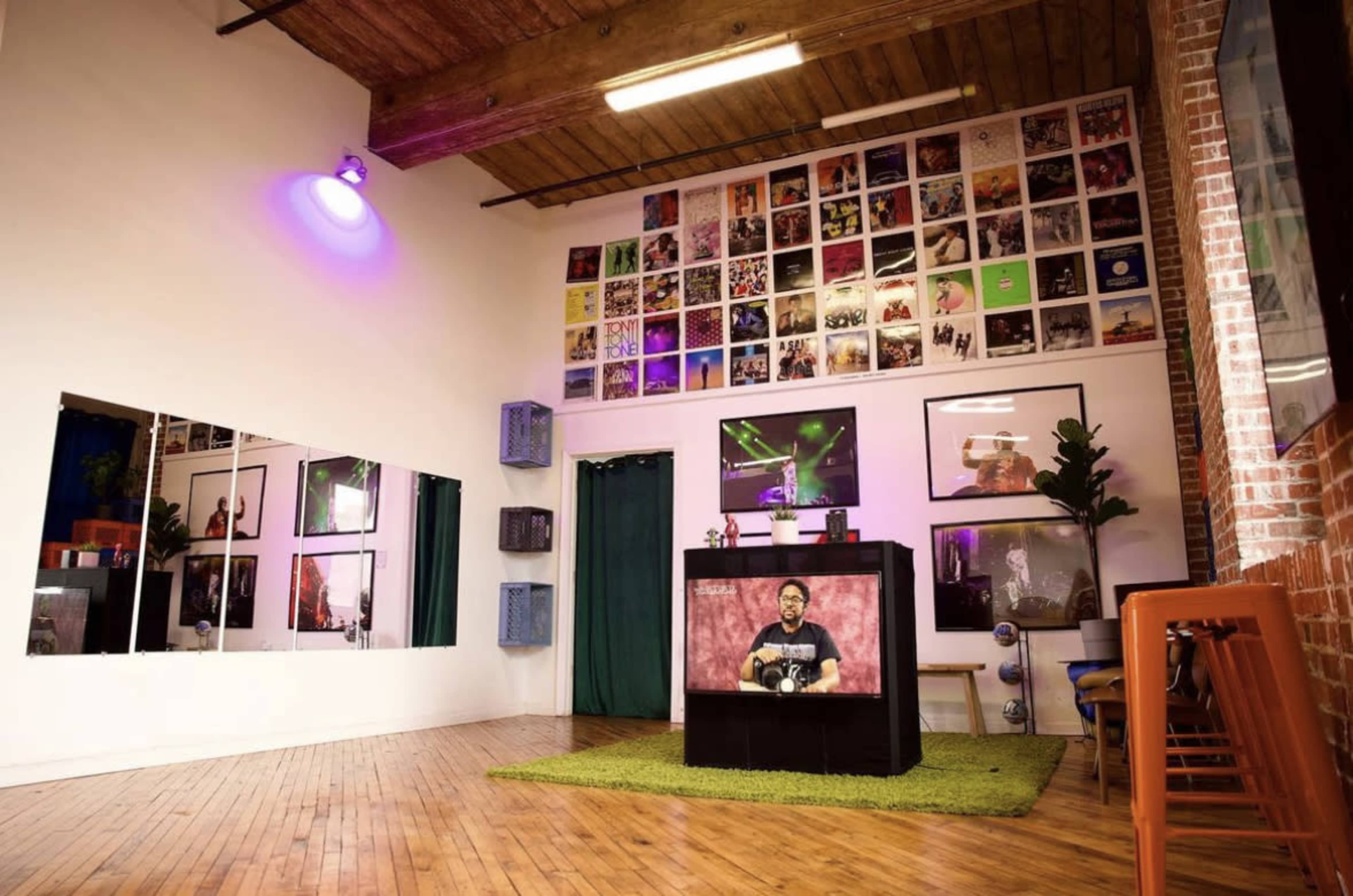 A brightly lit room with wooden floors, a wall covered in framed photographs and posters, a dark display table at the center, and large mirrors on one side.