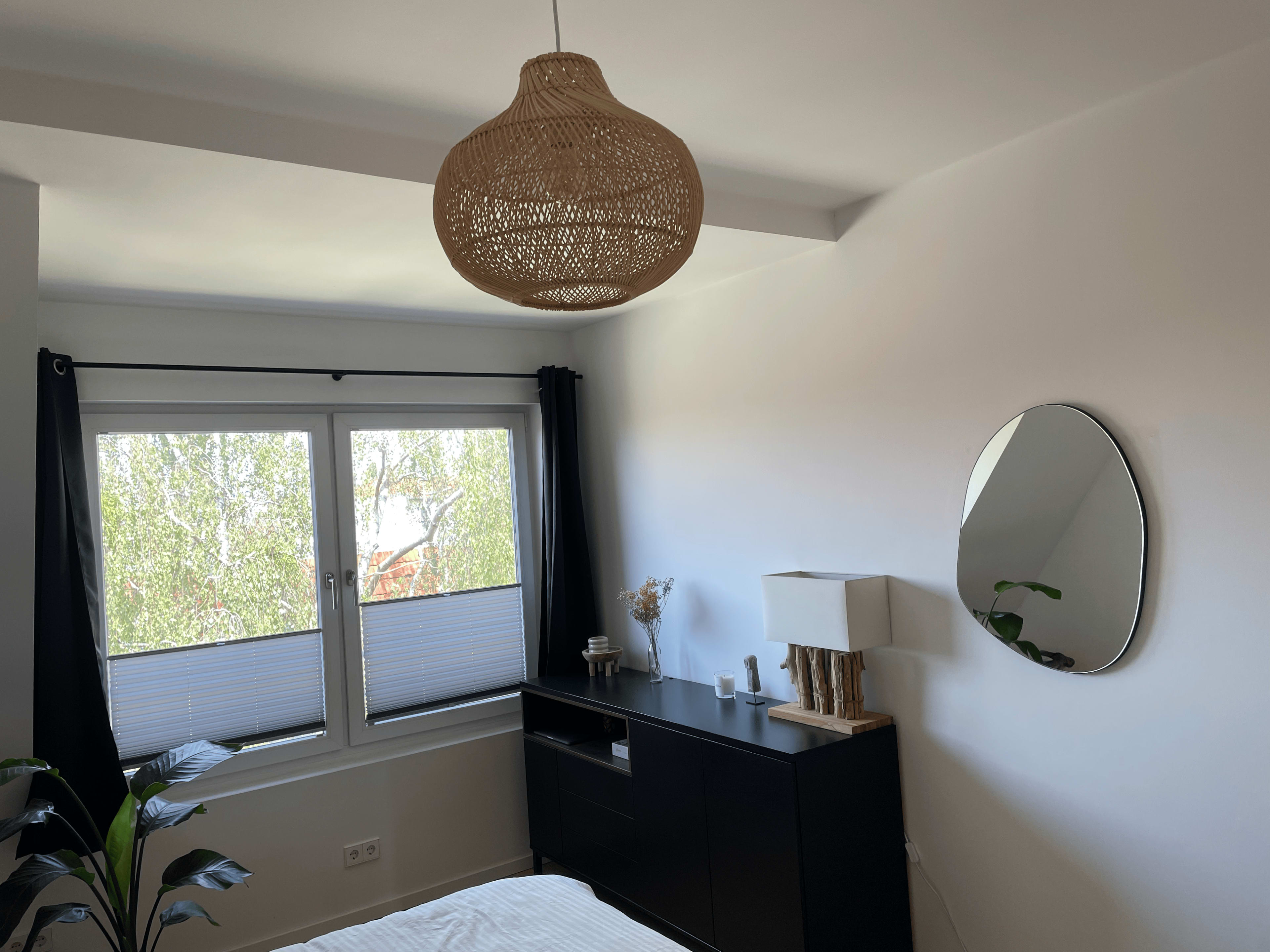 A bright bedroom featuring a black dresser, a round mirror, a lamp, and a window with light-colored blinds.