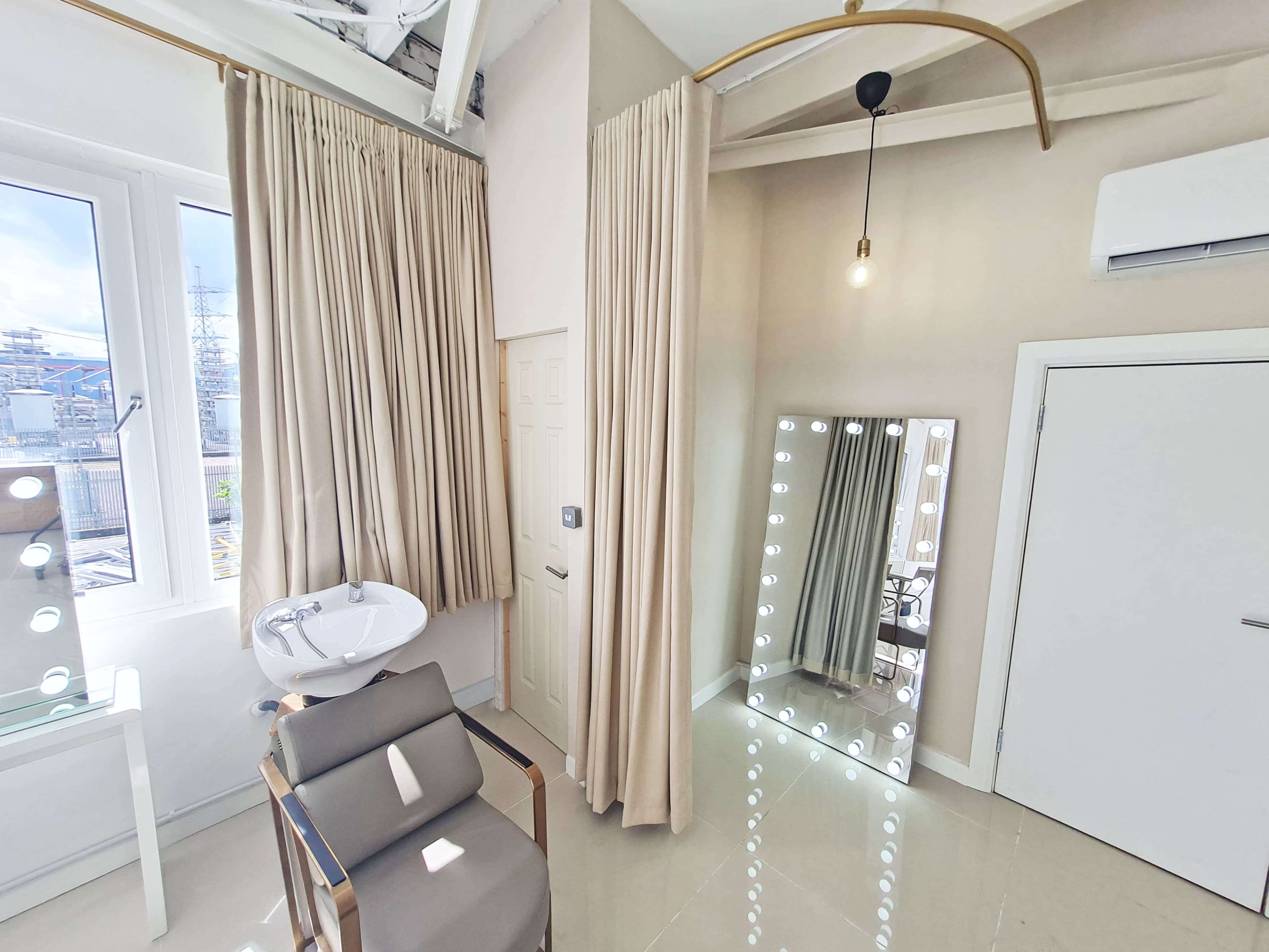 A well-lit room featuring a mirror with bright lights, a chair, a wall-mounted sink, and curtains covering a window.