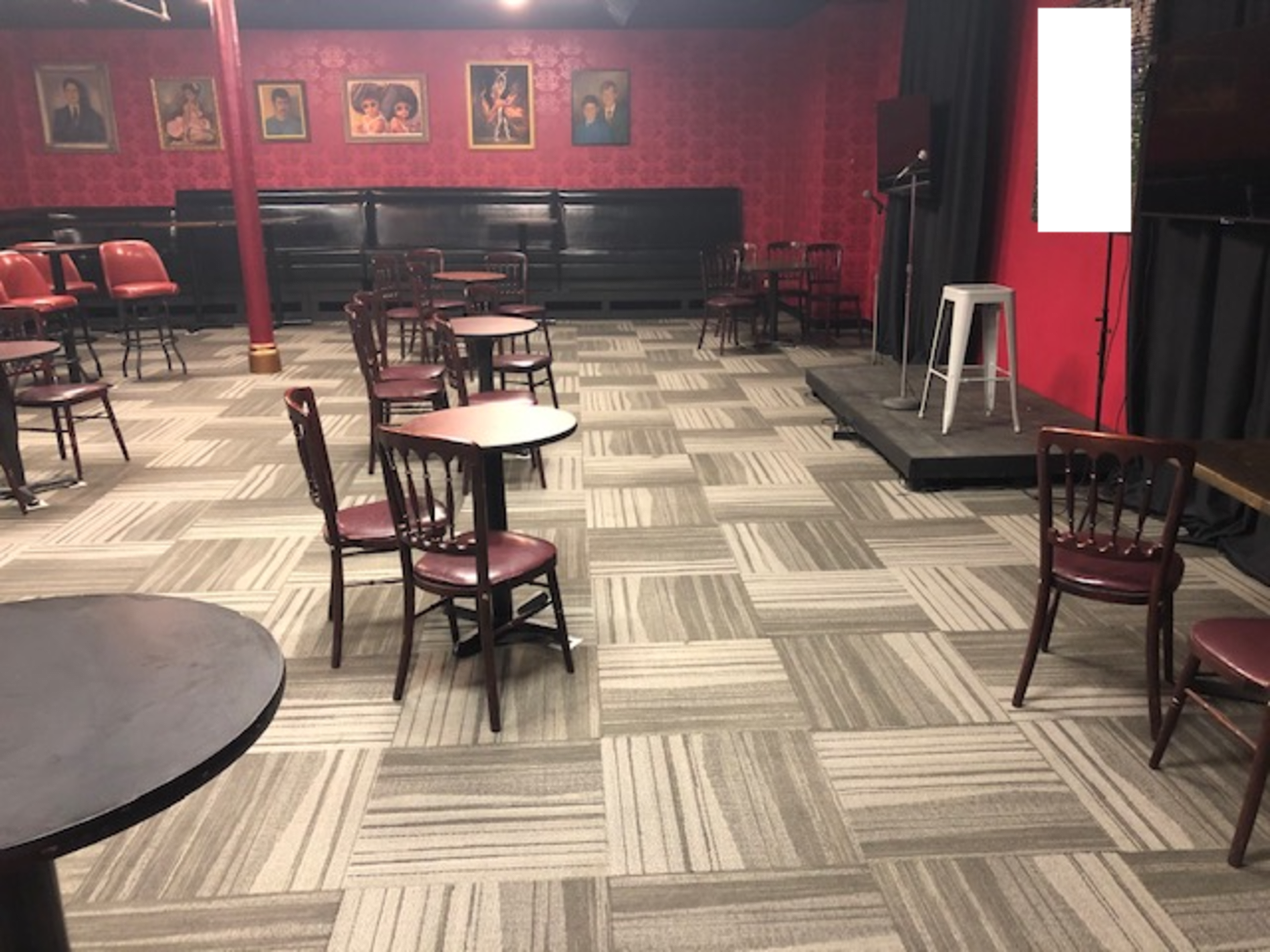 A dimly lit venue with a patterned carpet, several tables, and chairs arranged around a small stage.