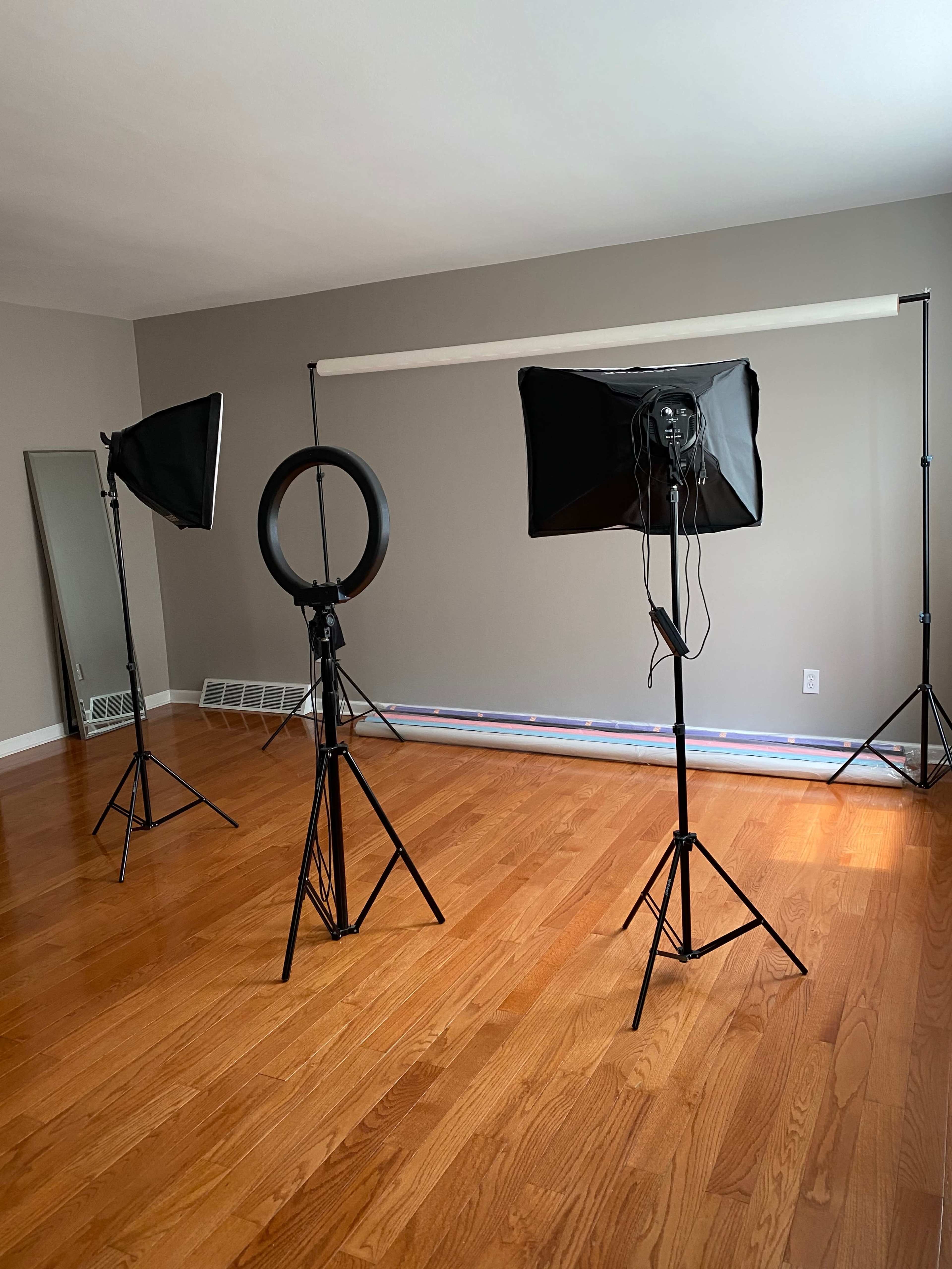 A photography studio setup with three lighting stands, including a ring light, arranged in a room with wooden flooring and a large mirror.