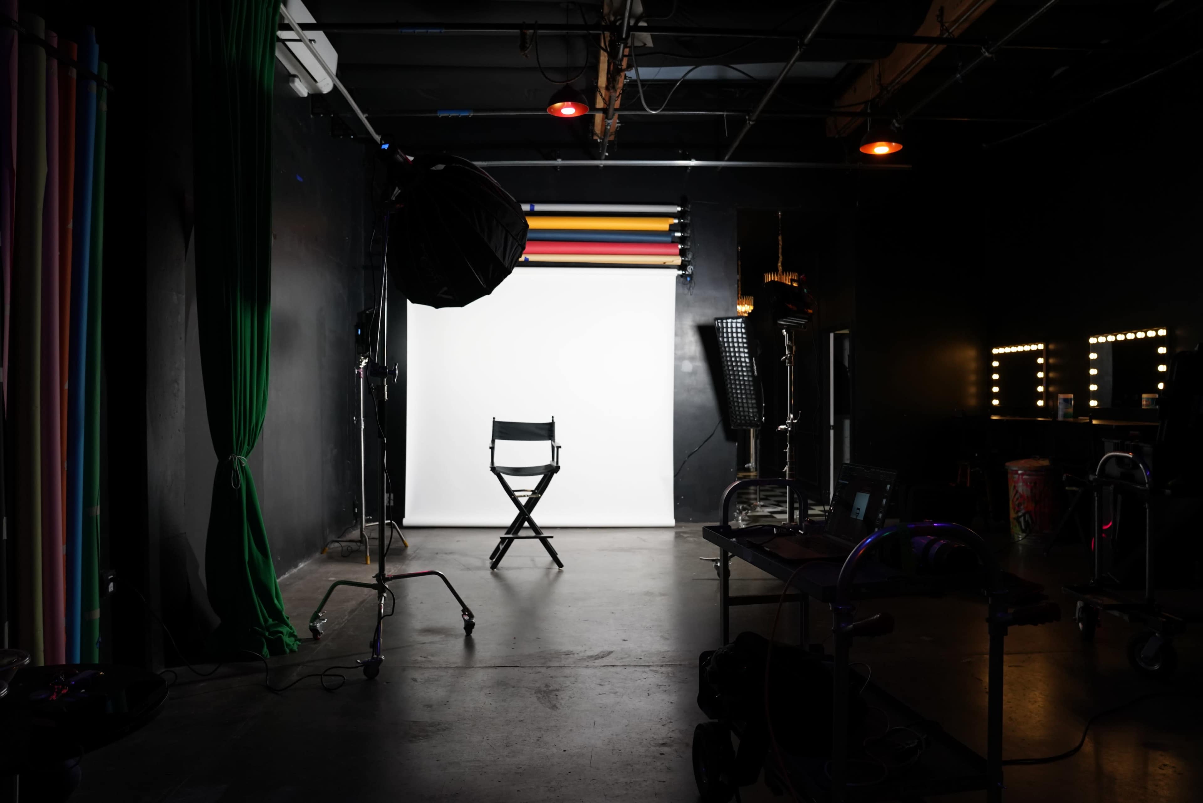 The image shows a photography studio with a black backdrop, a single director's chair centered in front of a white screen, and various lighting equipment arranged around the space.