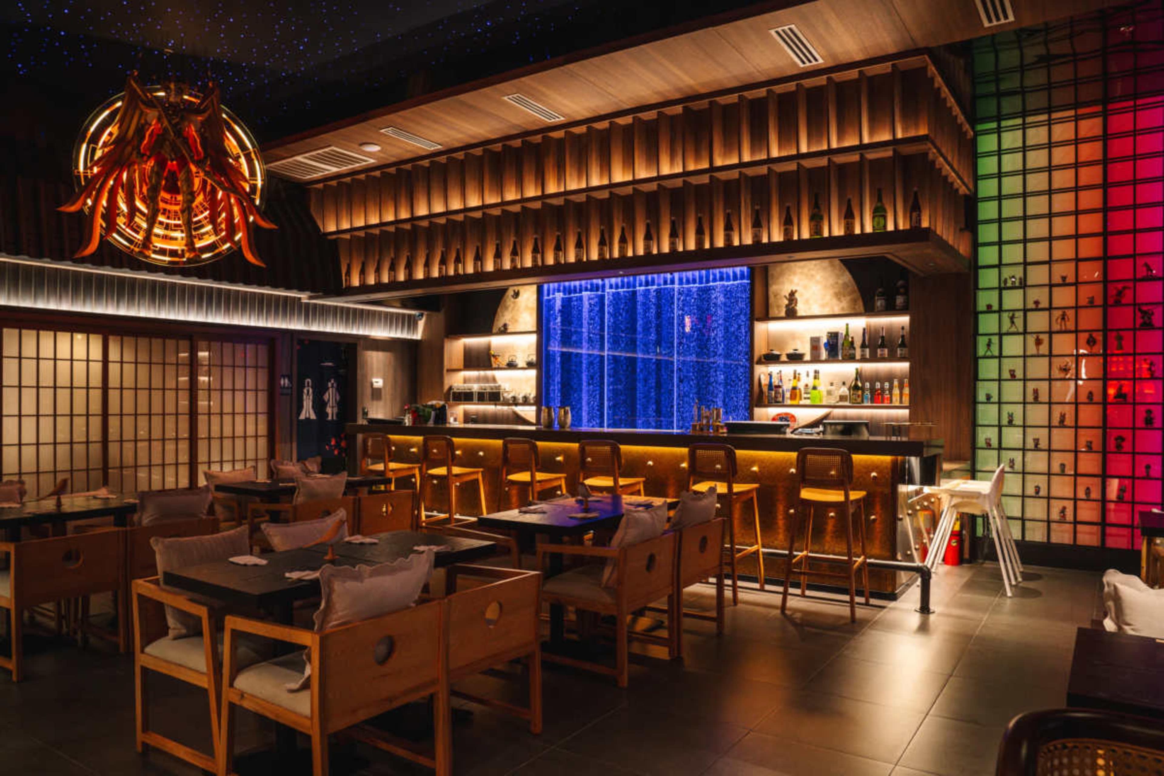 The image features a modern bar area with a colorful backlit display, wooden decor, and a seating arrangement that includes wooden chairs and tables.