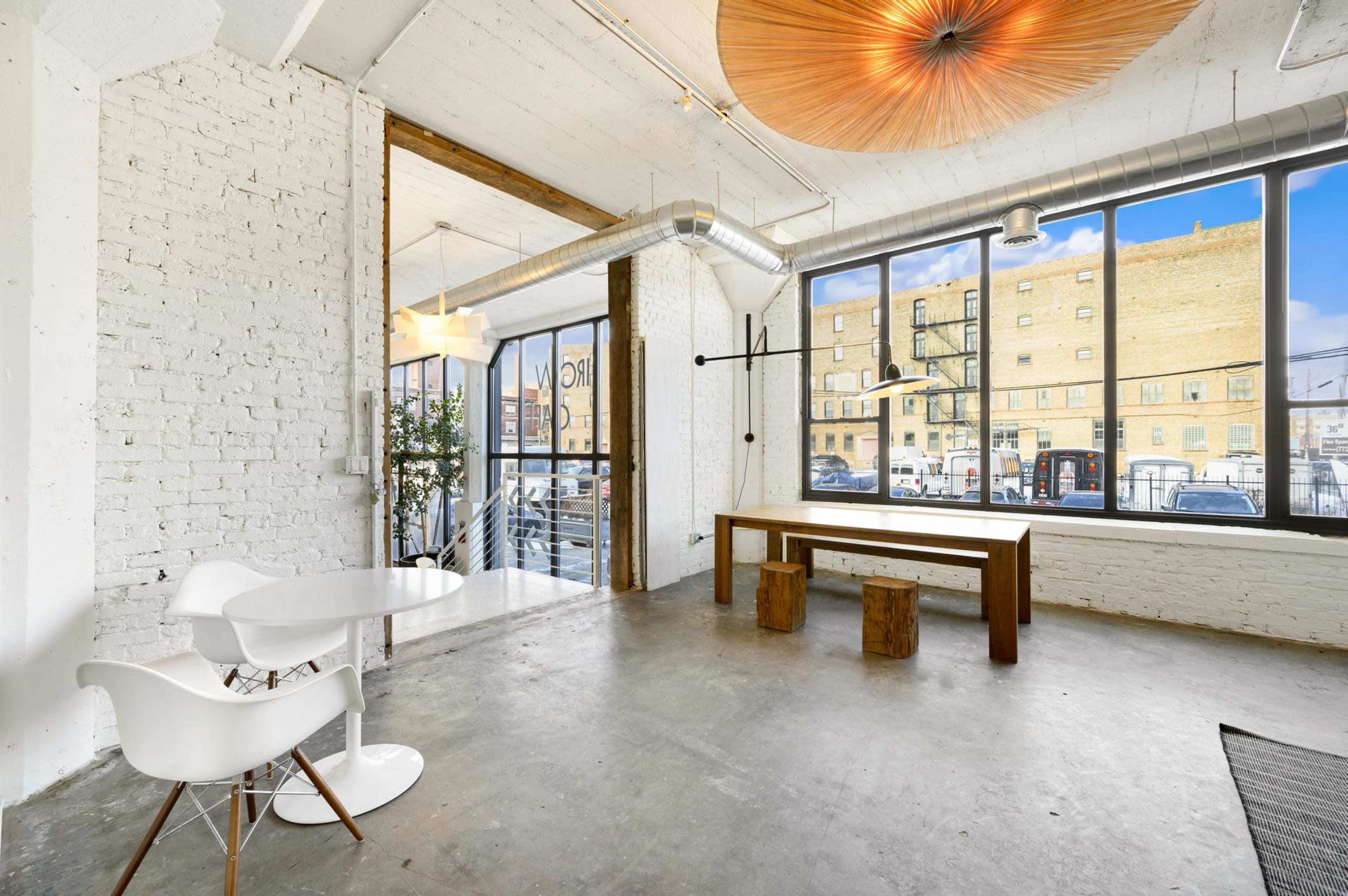 A bright industrial-style room features large windows, a wooden table, and minimalist furniture against a backdrop of exposed brick walls.