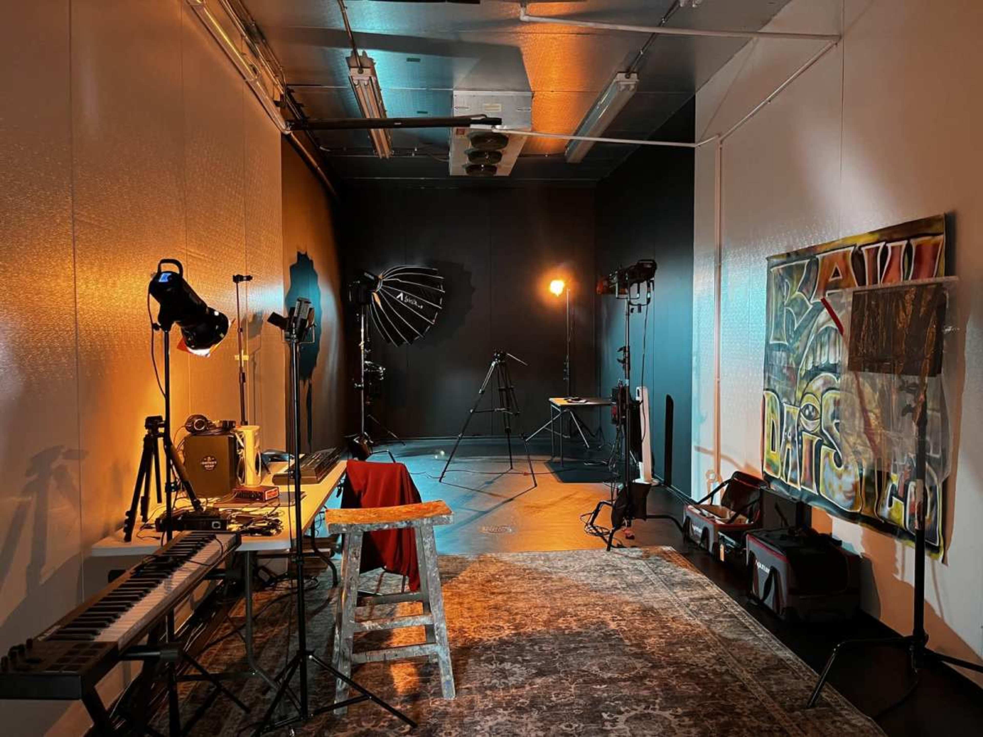 The image shows a studio space with various lighting equipment, a keyboard, a table, and an art piece on display against contrasting light and dark walls.