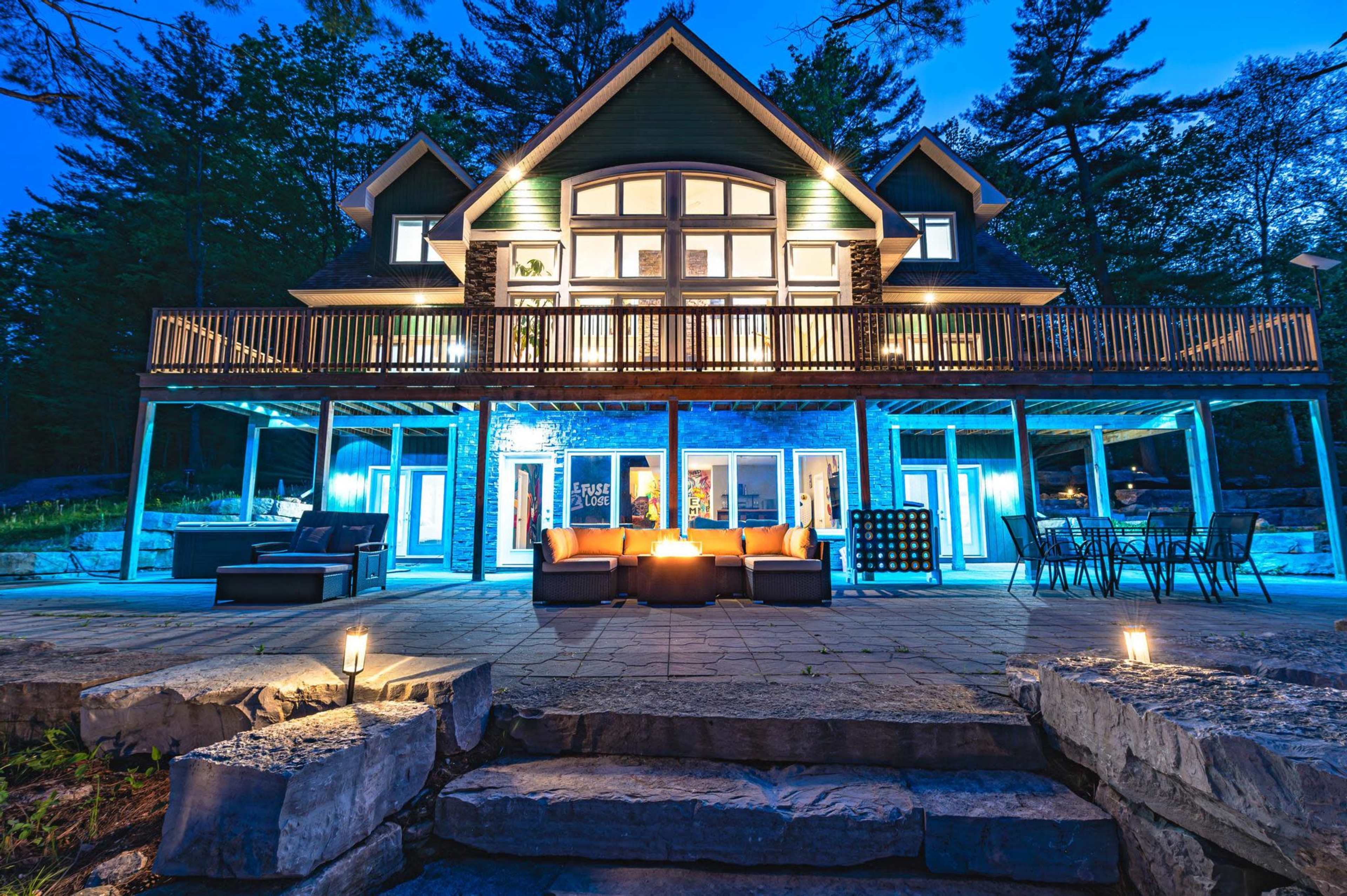 A large, modern house with a spacious deck is illuminated at dusk, featuring outdoor seating and a stone patio surrounded by trees.