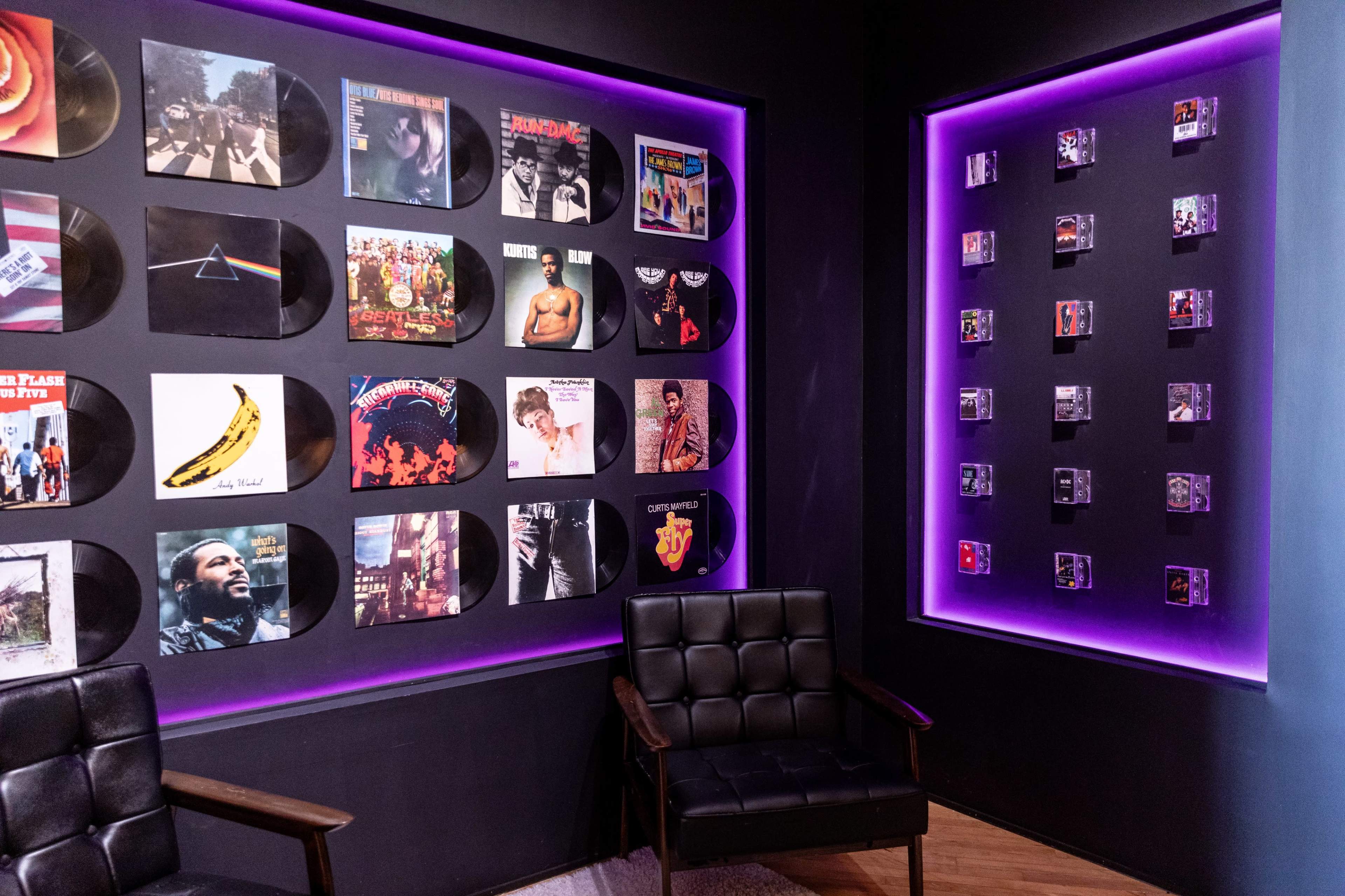 The image shows a music-themed room with two walls adorned with album covers and a pair of black chairs in the foreground.