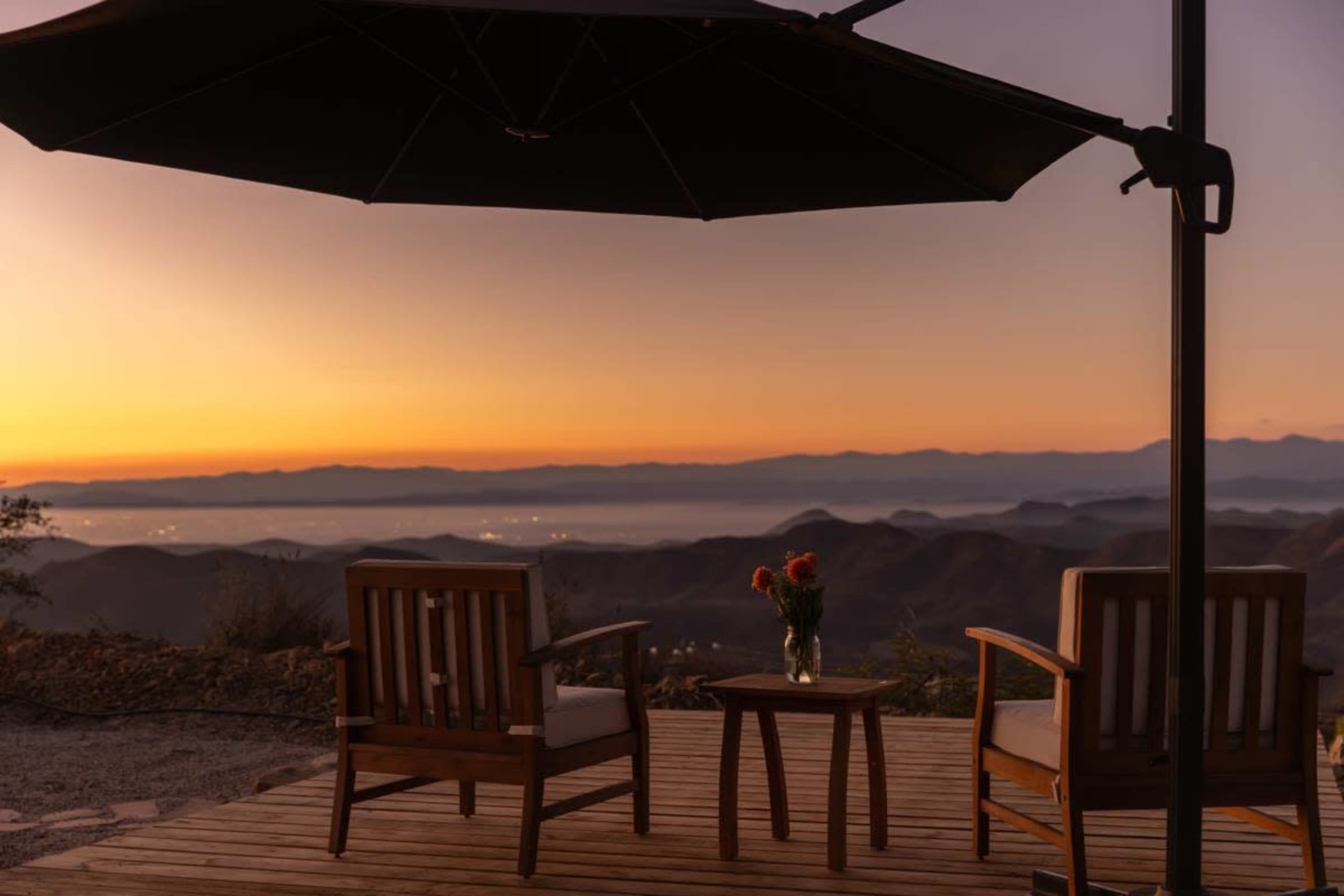 Two wooden chairs and a small table with a vase of flowers sit on a deck overlooking mountainous terrain at sunset.