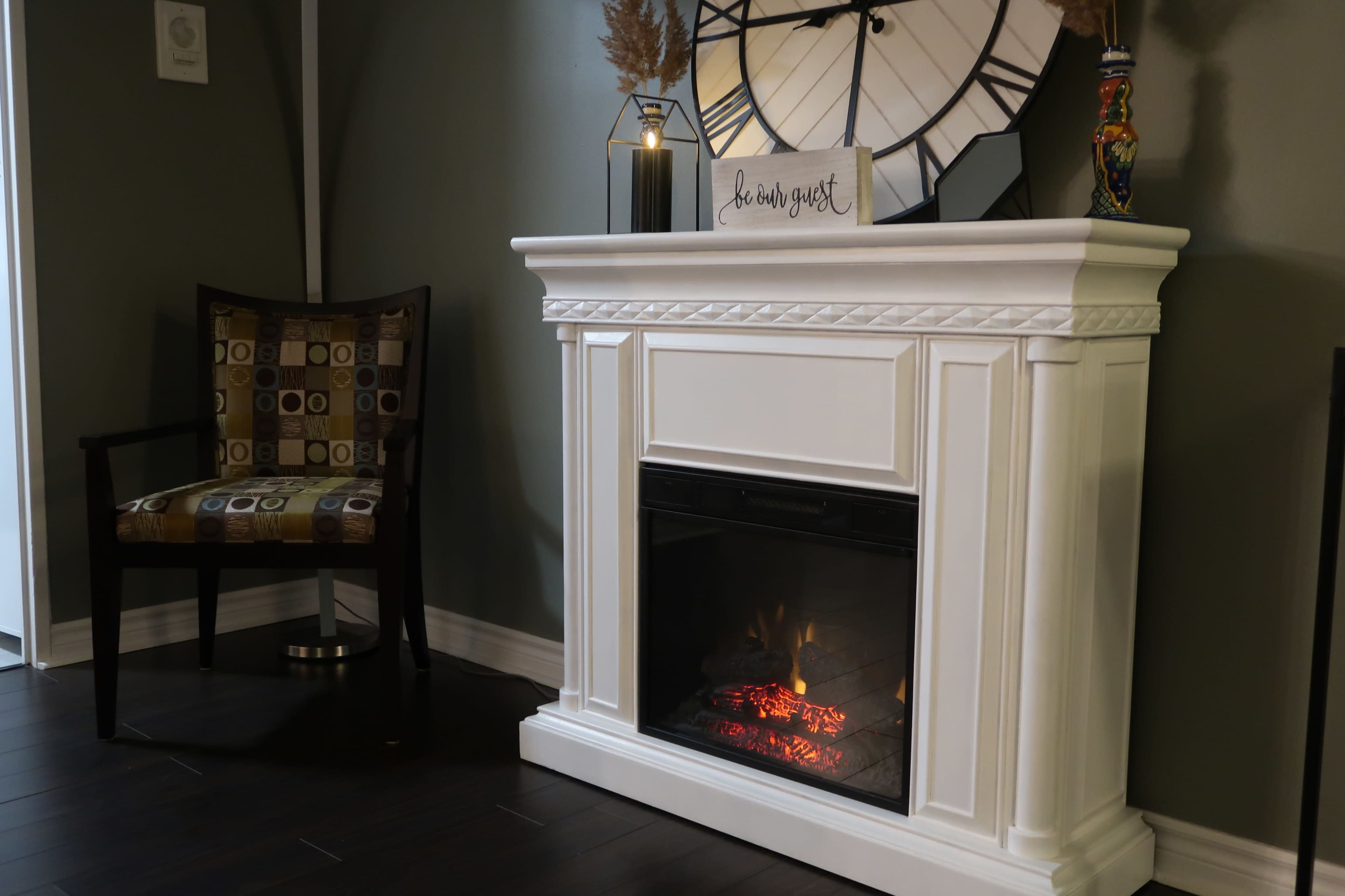 A white fireplace with an electric flame feature, a decorative clock on the mantle, and a patterned chair nearby in a cozy room.