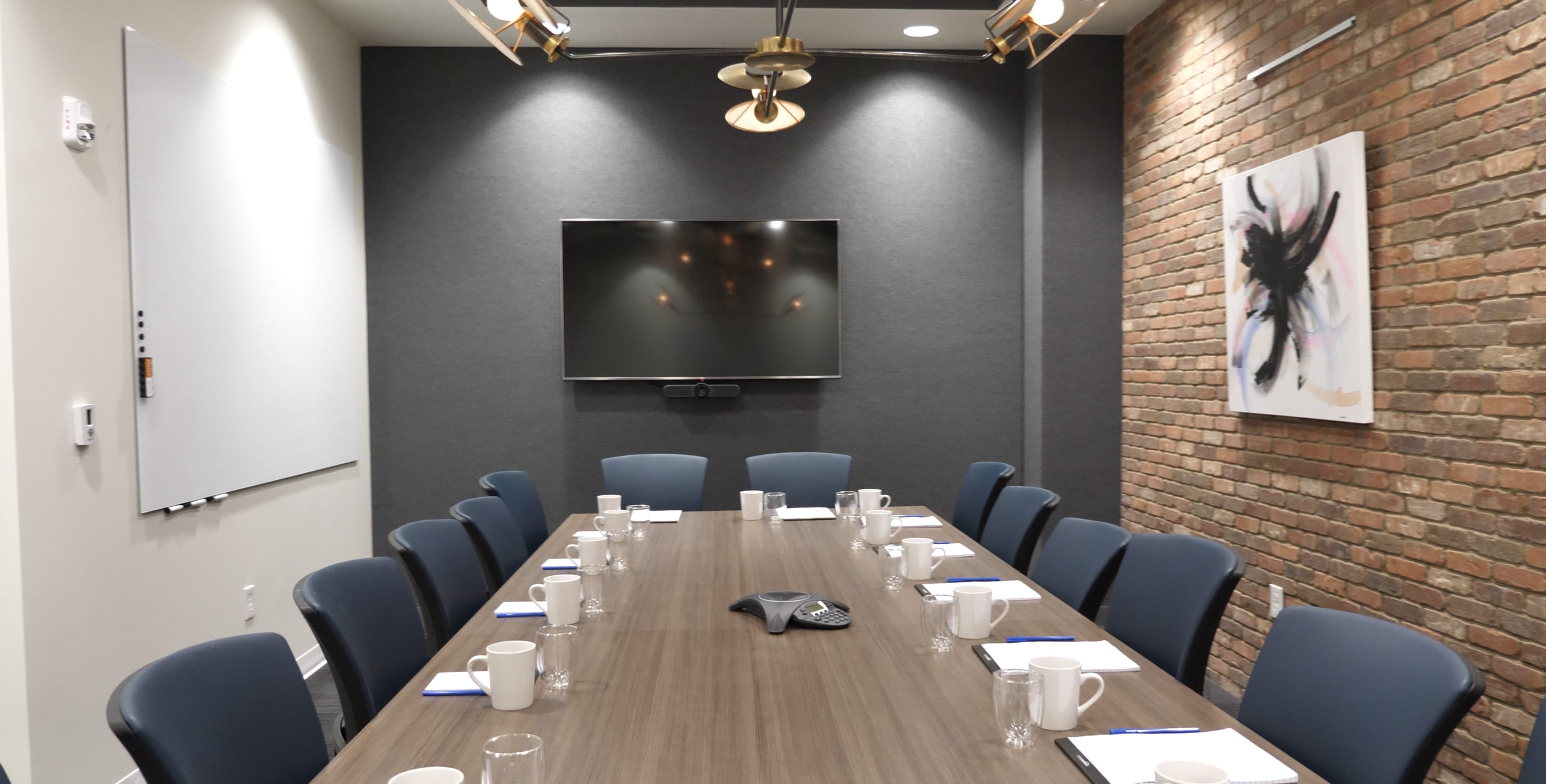 A modern conference room features a long wooden table with chairs, a large wall-mounted screen, a whiteboard, and brick accents.