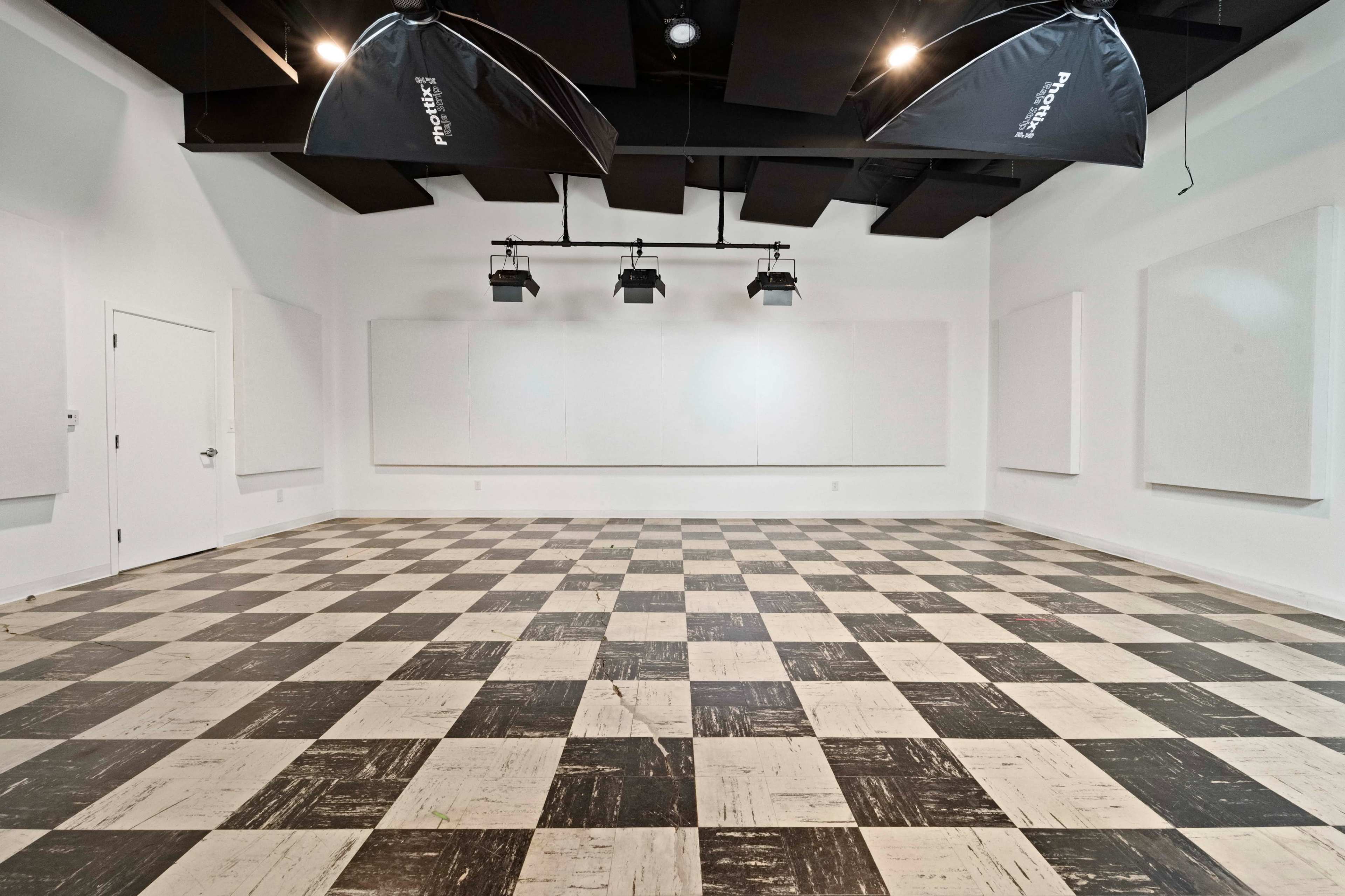 The image shows a spacious, empty room with a checkered floor, soundproofing panels on the walls, and studio lights hanging from the ceiling.