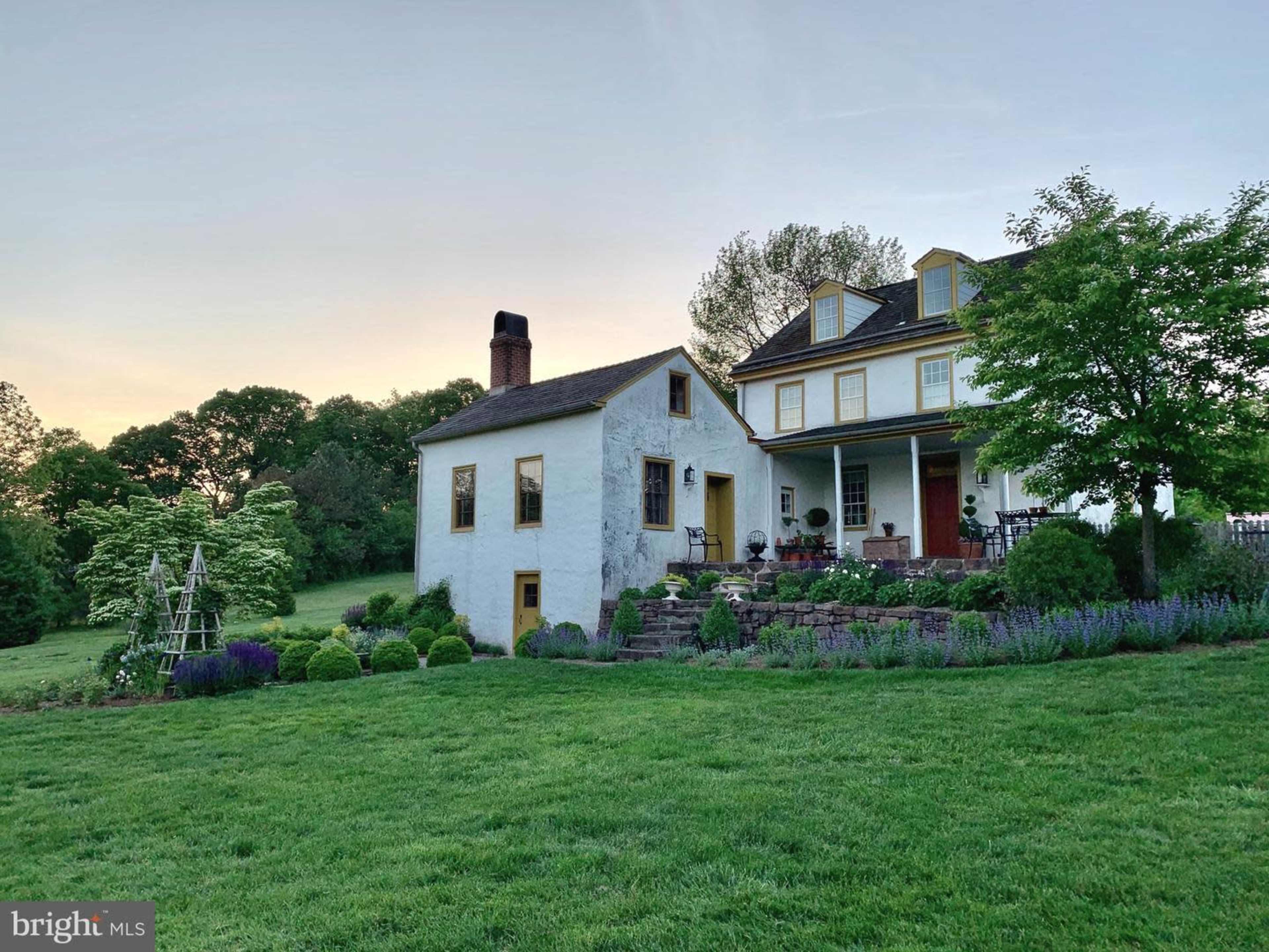 A large two-story house with yellow accents stands next to a smaller, white structure, surrounded by a neatly landscaped yard featuring green grass and garden beds.