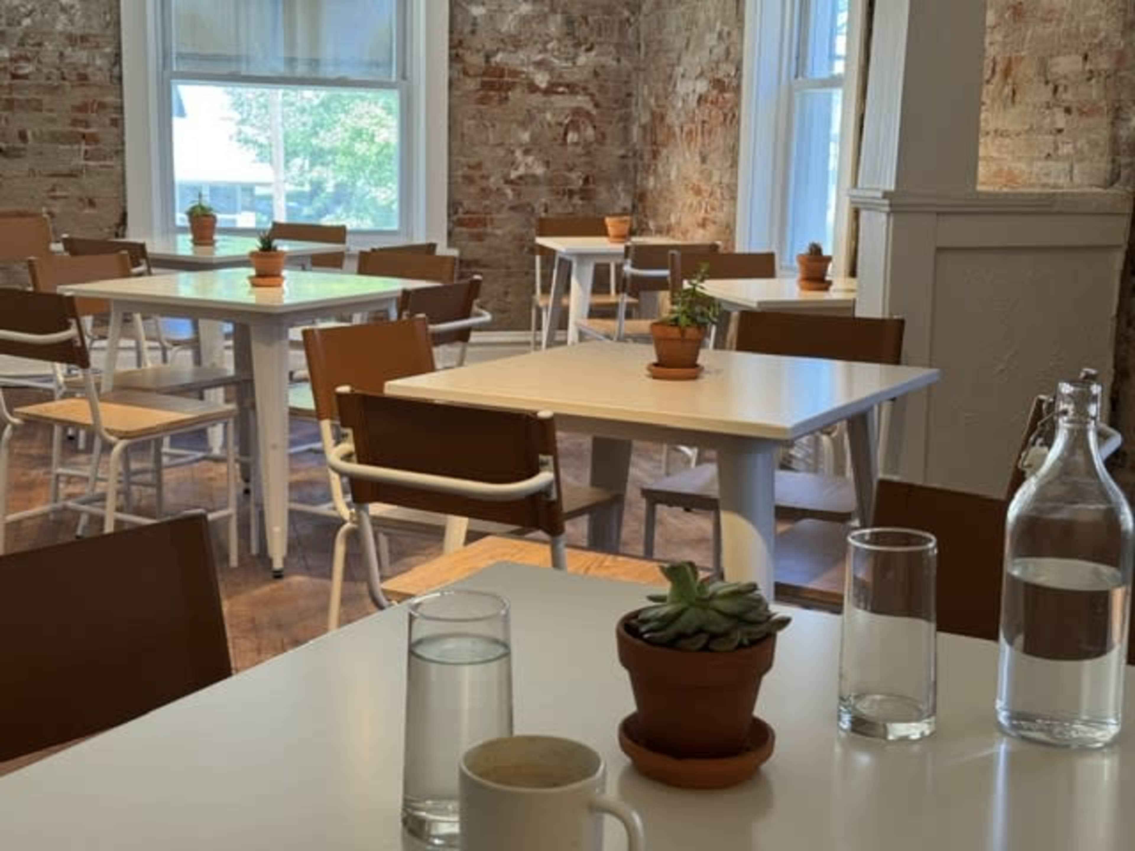The image shows a minimalist café interior with several wooden tables and chairs, each adorned with small potted plants.