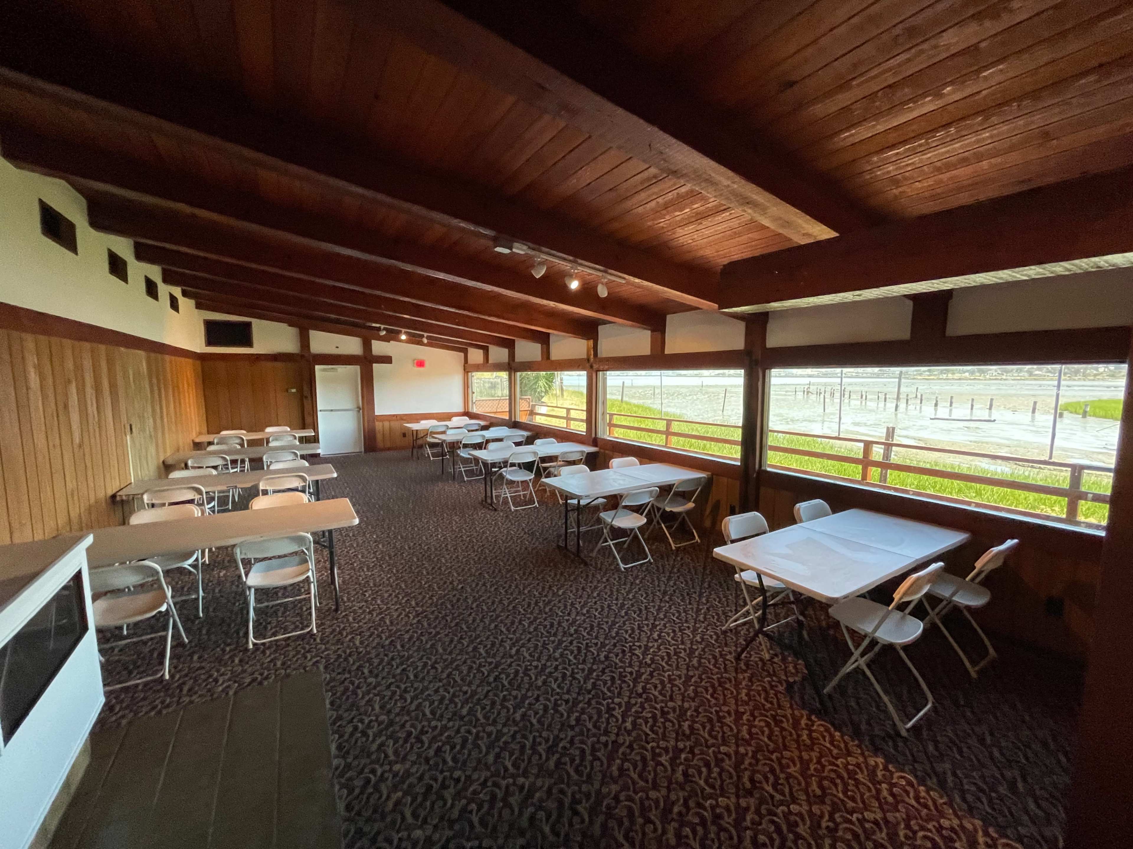 A spacious room with wooden beams, tables arranged in rows, and large windows overlooking a grassy area.