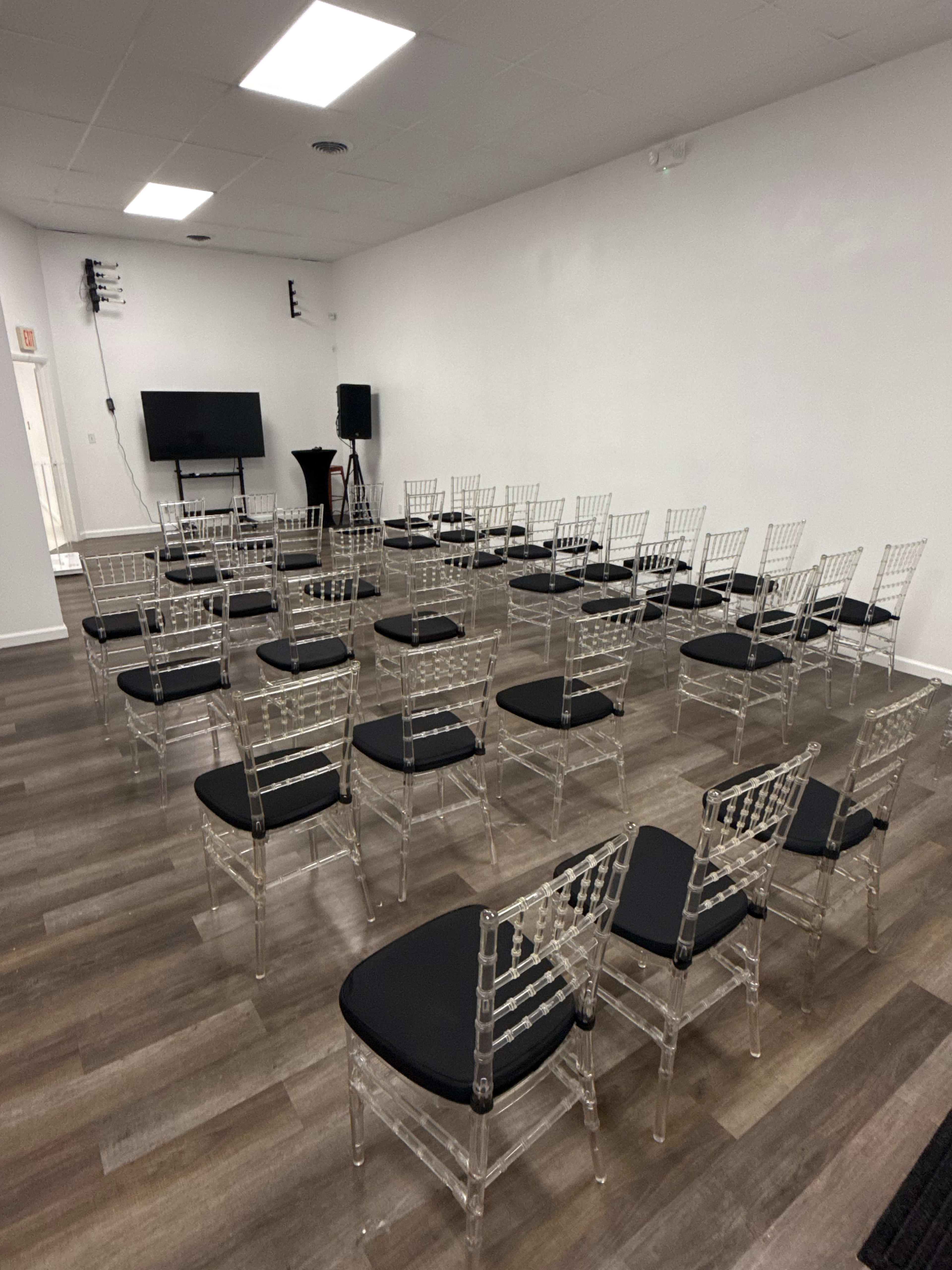 The image shows a room set up with several rows of transparent chairs with black cushions arranged for a gathering or event.