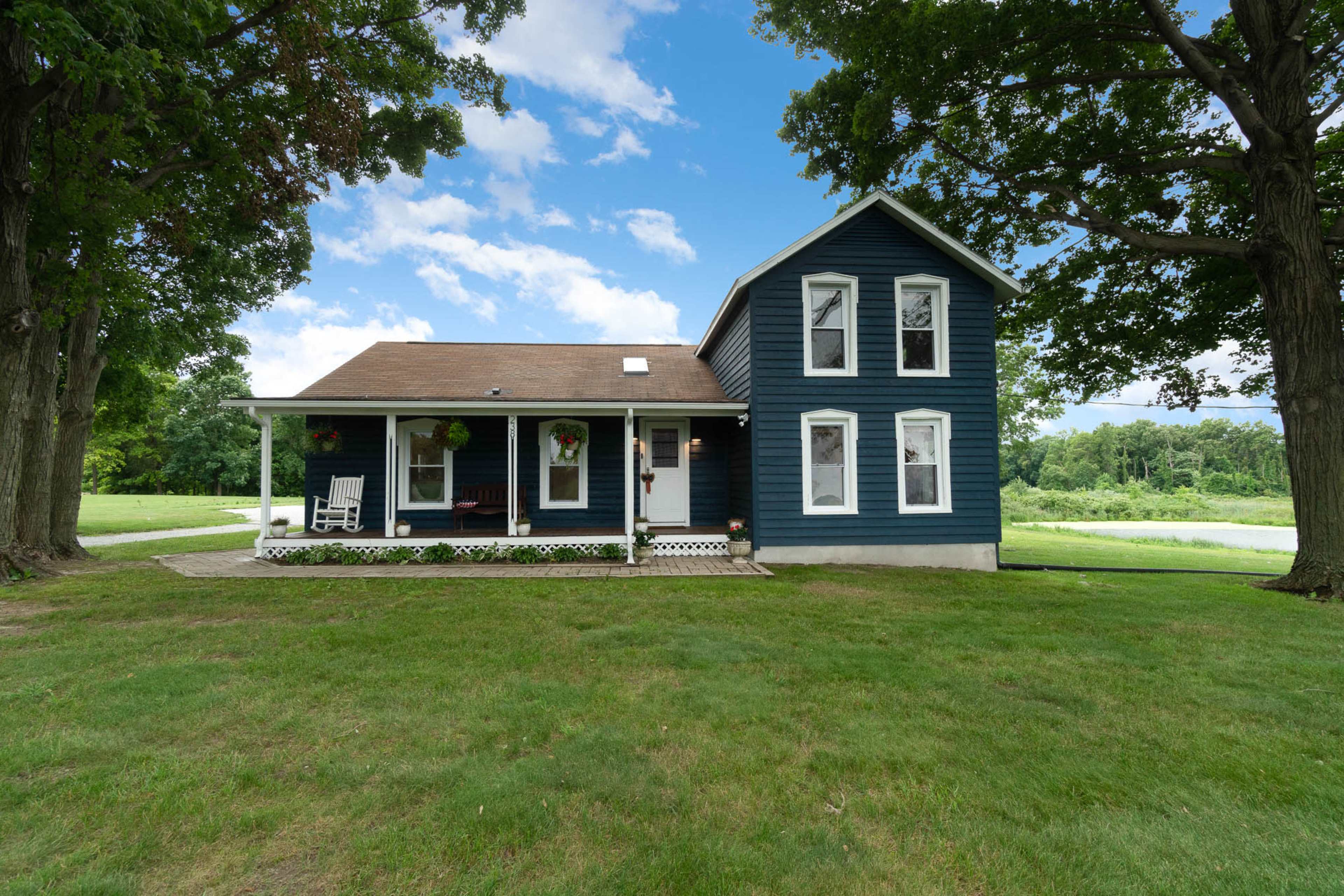 A blue two-story house with a porch is situated on a grassy lawn surrounded by trees.