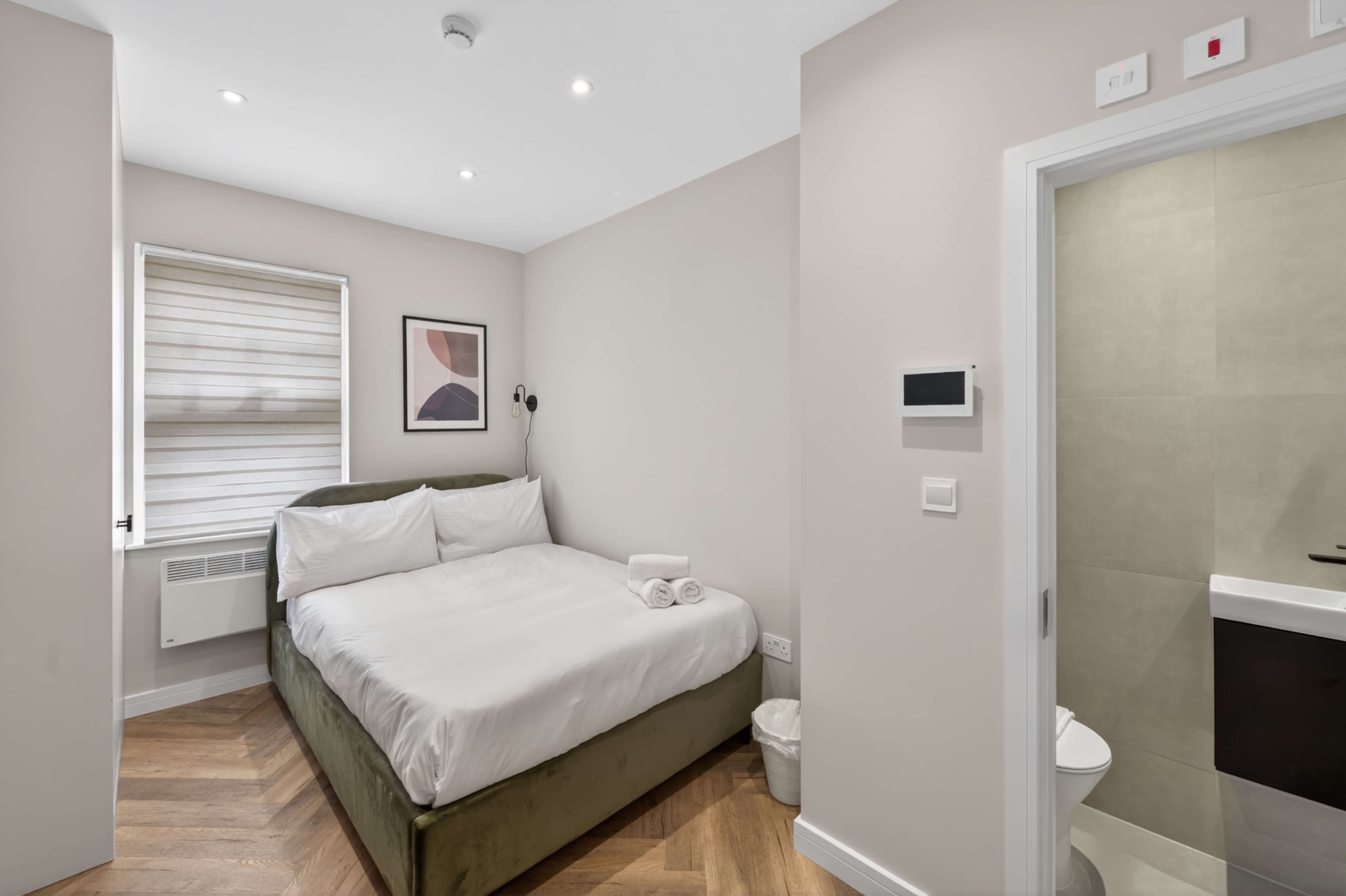 The image shows a small hotel room with a double bed, a window with shades, a minimalist art piece on the wall, and an adjoining bathroom.