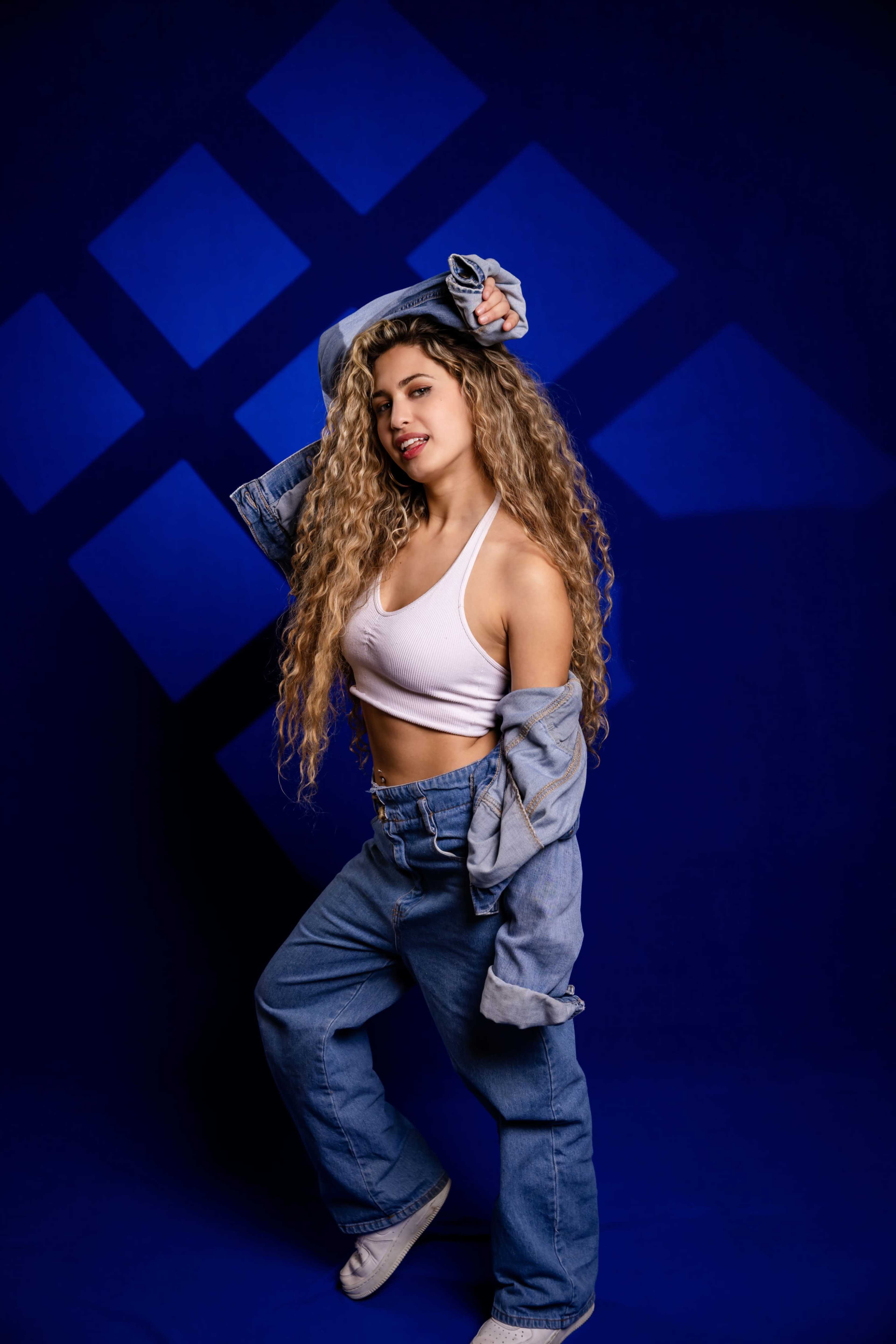 A person with long, curly hair poses in a white crop top and oversized denim jacket against a blue backdrop with geometric shapes.