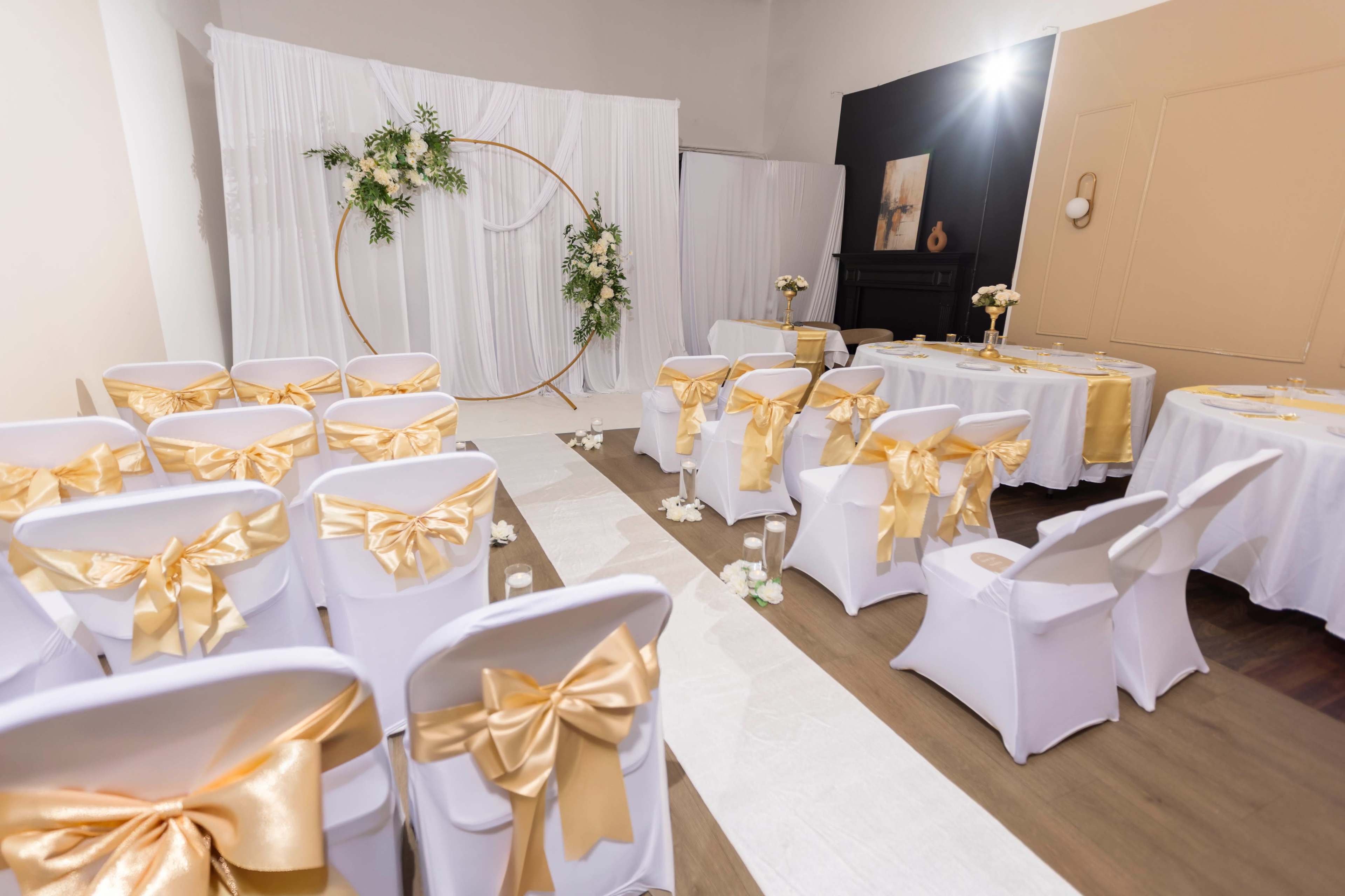 The image shows a decorated event space featuring rows of white chairs with gold bow ties, a circular floral backdrop, and tables set with white tablecloths.