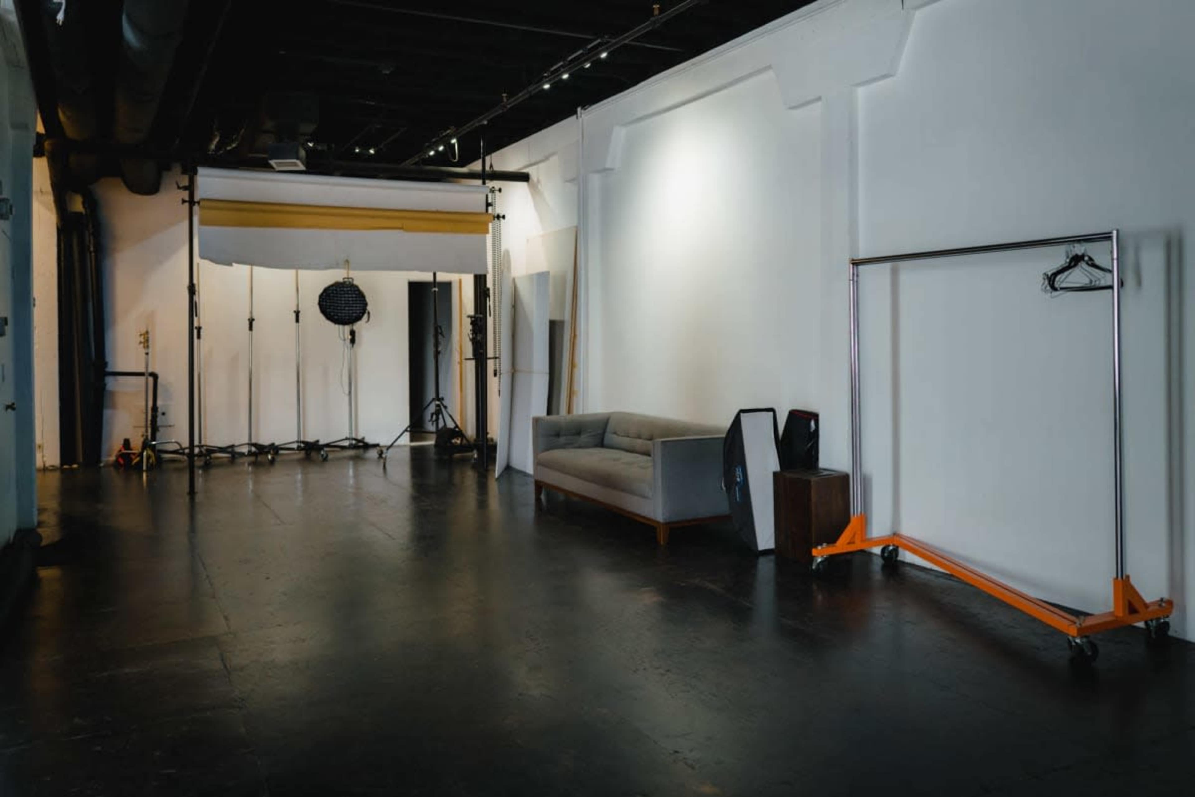 The image shows a spacious studio with a gray couch, a clothing rack on wheels, and various lighting equipment set up against a stark white wall.