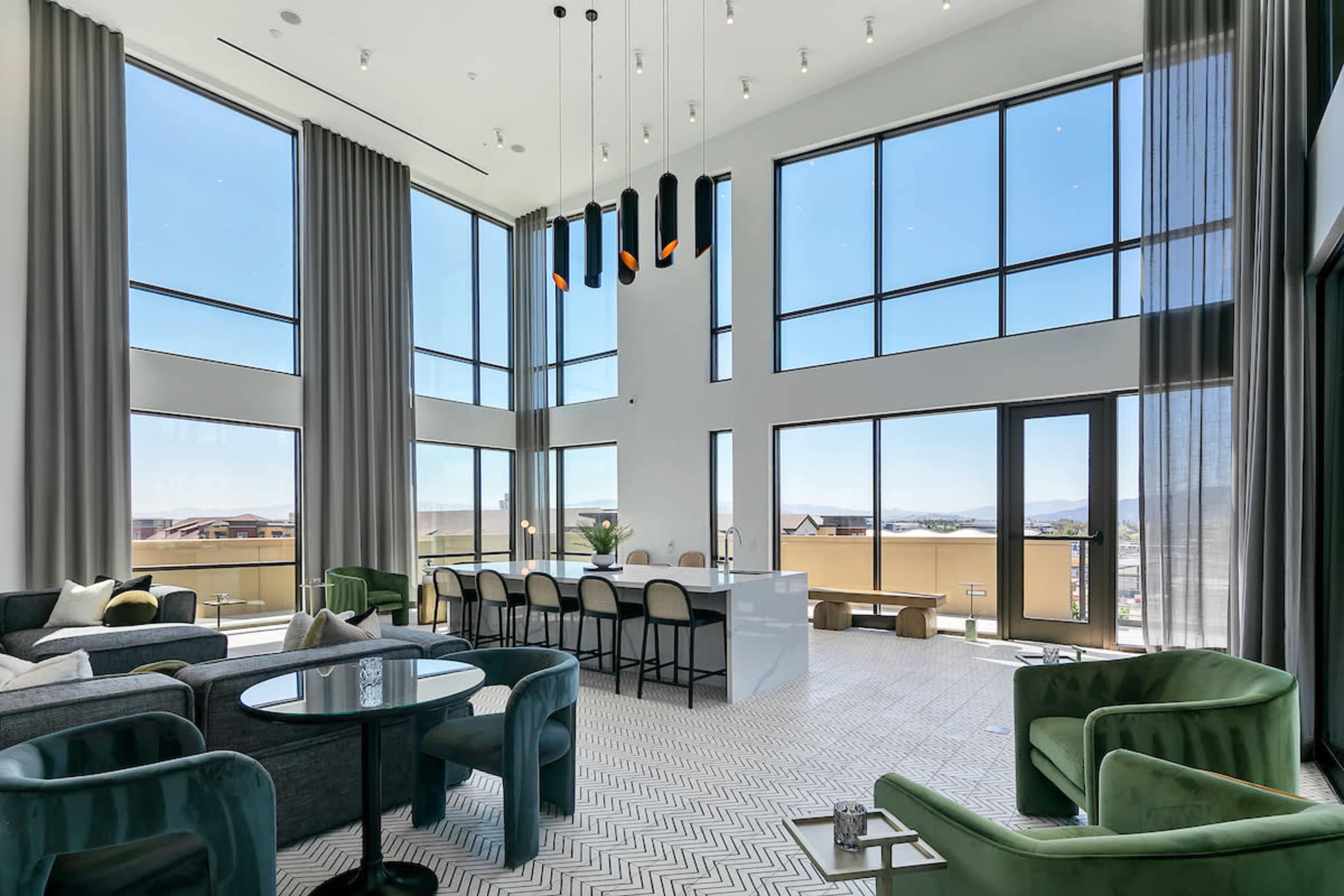 The image shows a modern living space with large windows, high ceilings, and a kitchen area featuring a white countertop and bar seating.