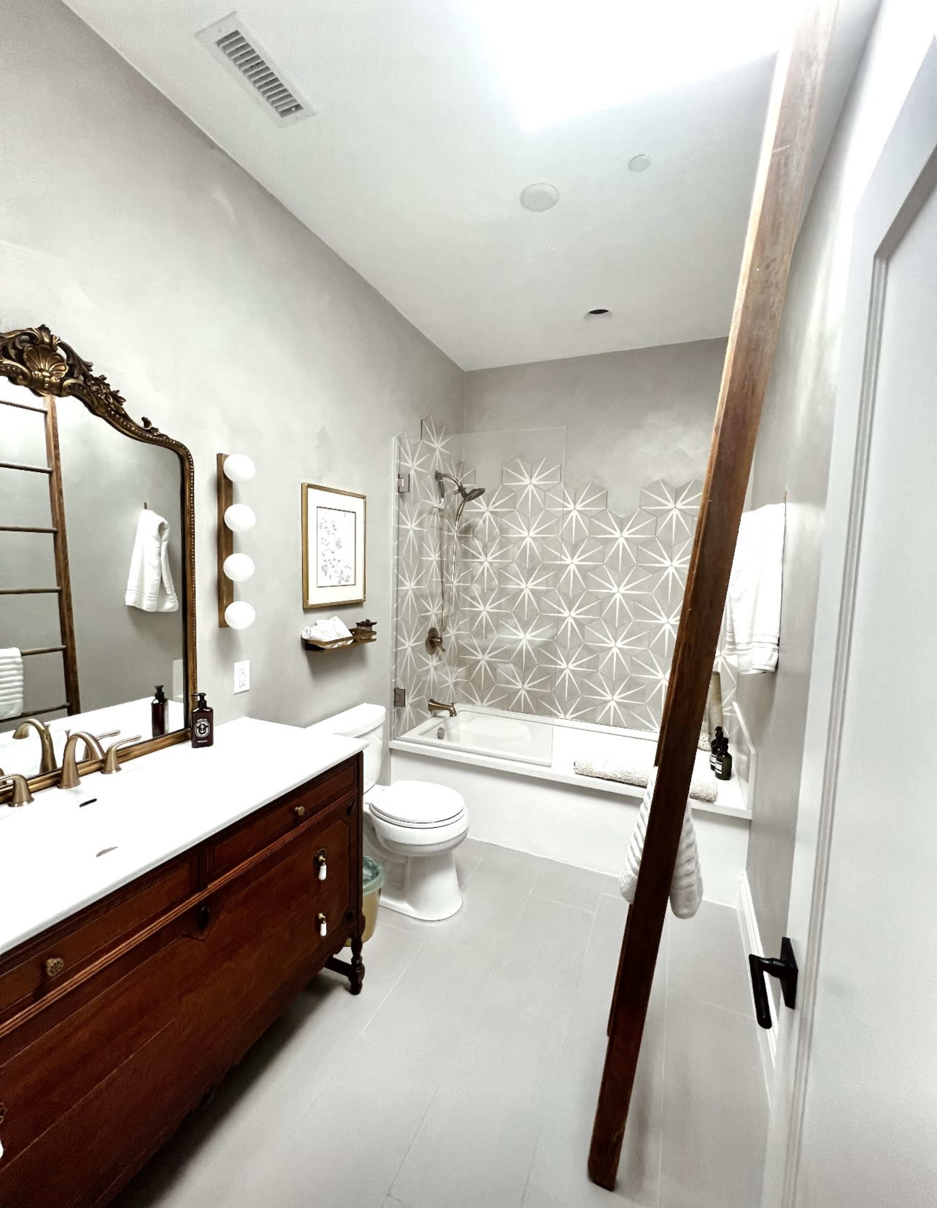 The bathroom features a modern design with a wooden vanity, a white bathtub surrounded by patterned tiles, and a large mirror on one wall.