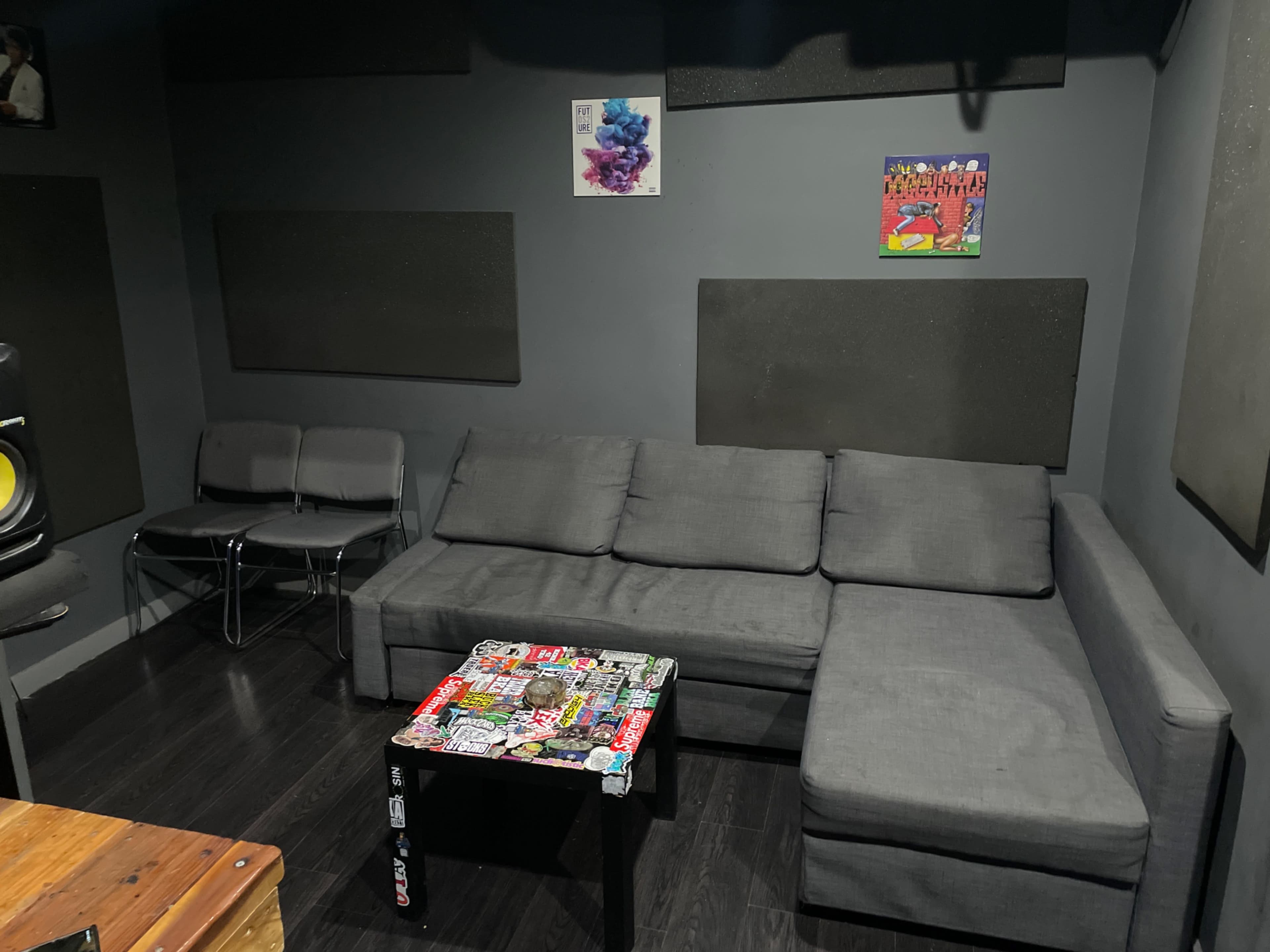 The image shows a small music studio with a gray sectional sofa, a black table covered in stickers, and soundproofing panels on the walls.
