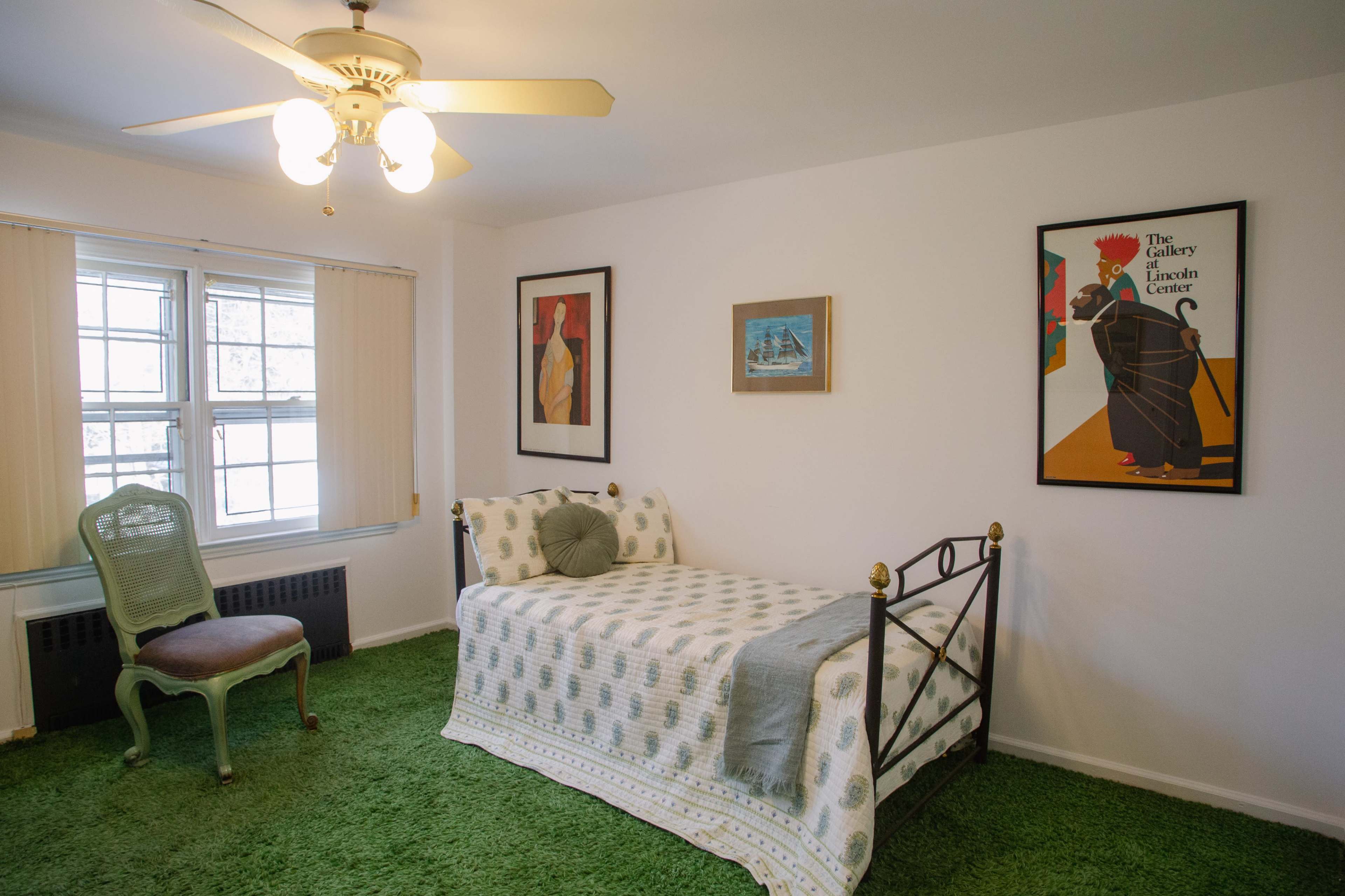 A bedroom features a single bed with a patterned quilt, a green plush carpet, a chair, and framed artworks on the walls.