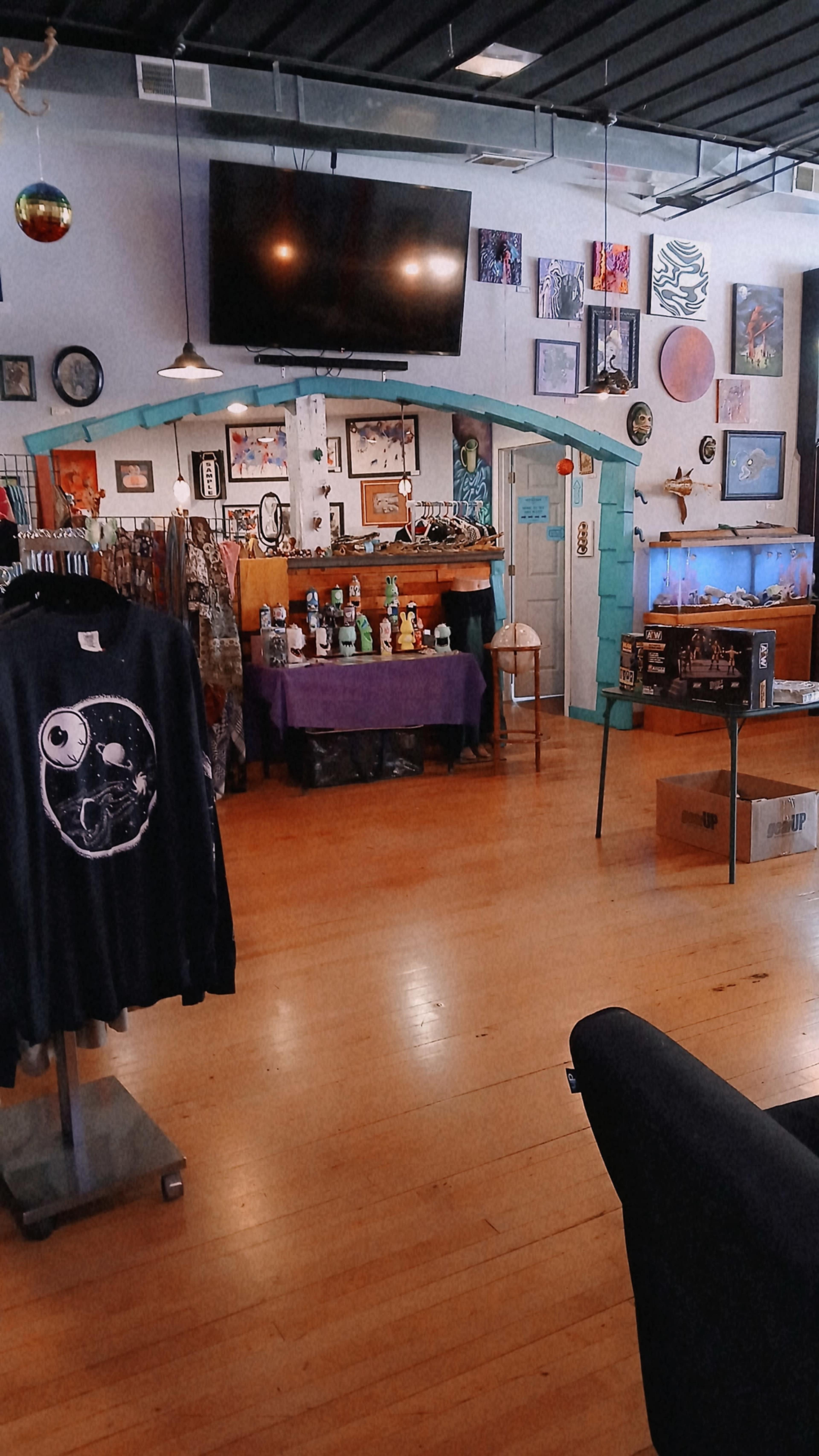 The image shows an interior view of a shop featuring displayed clothing, a table with various items, and art pieces adorning the walls.