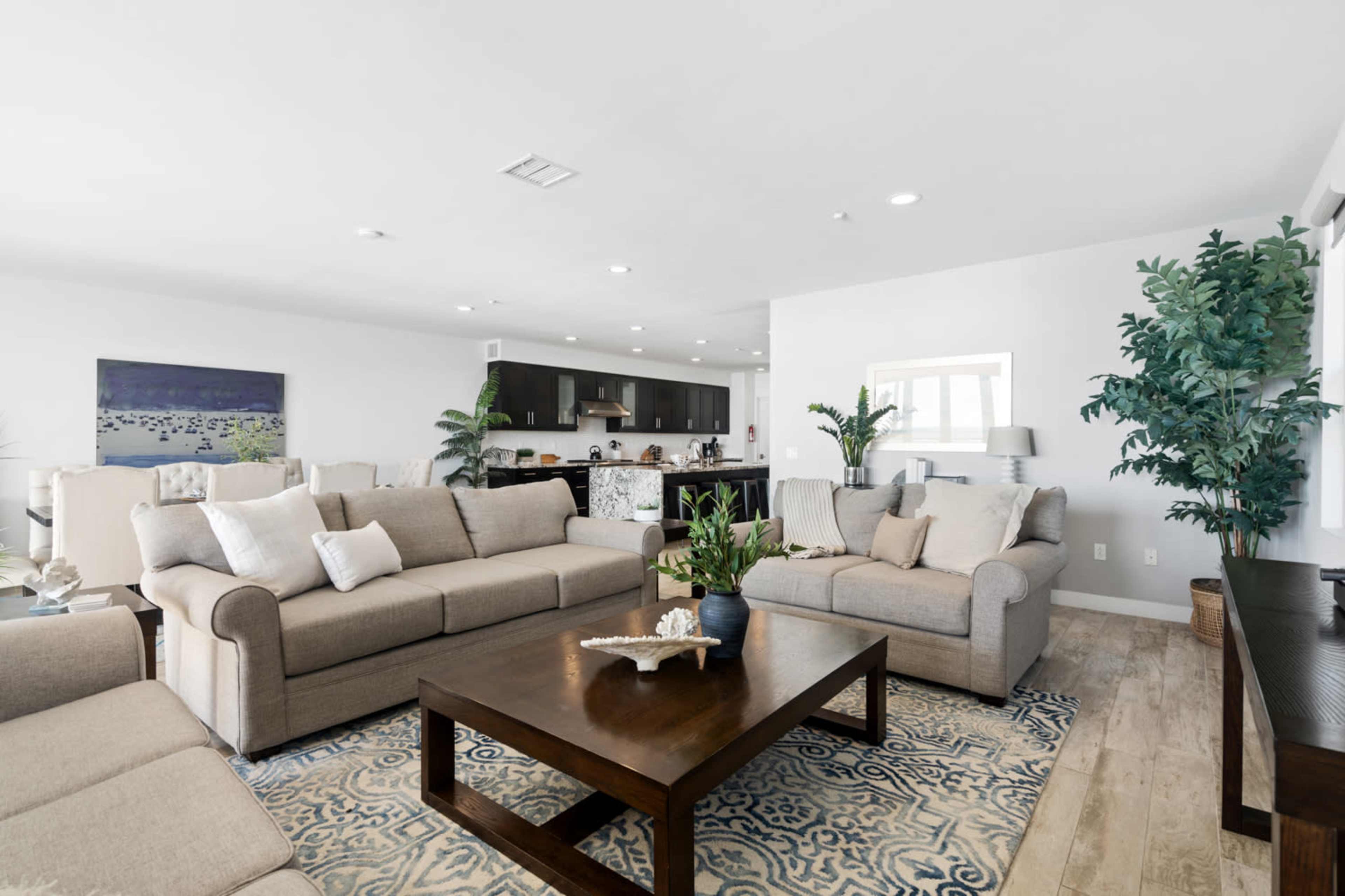 A modern living room with two light-colored sofas, a large wooden coffee table, and a view into a kitchen area with dark cabinetry and a marble countertop.