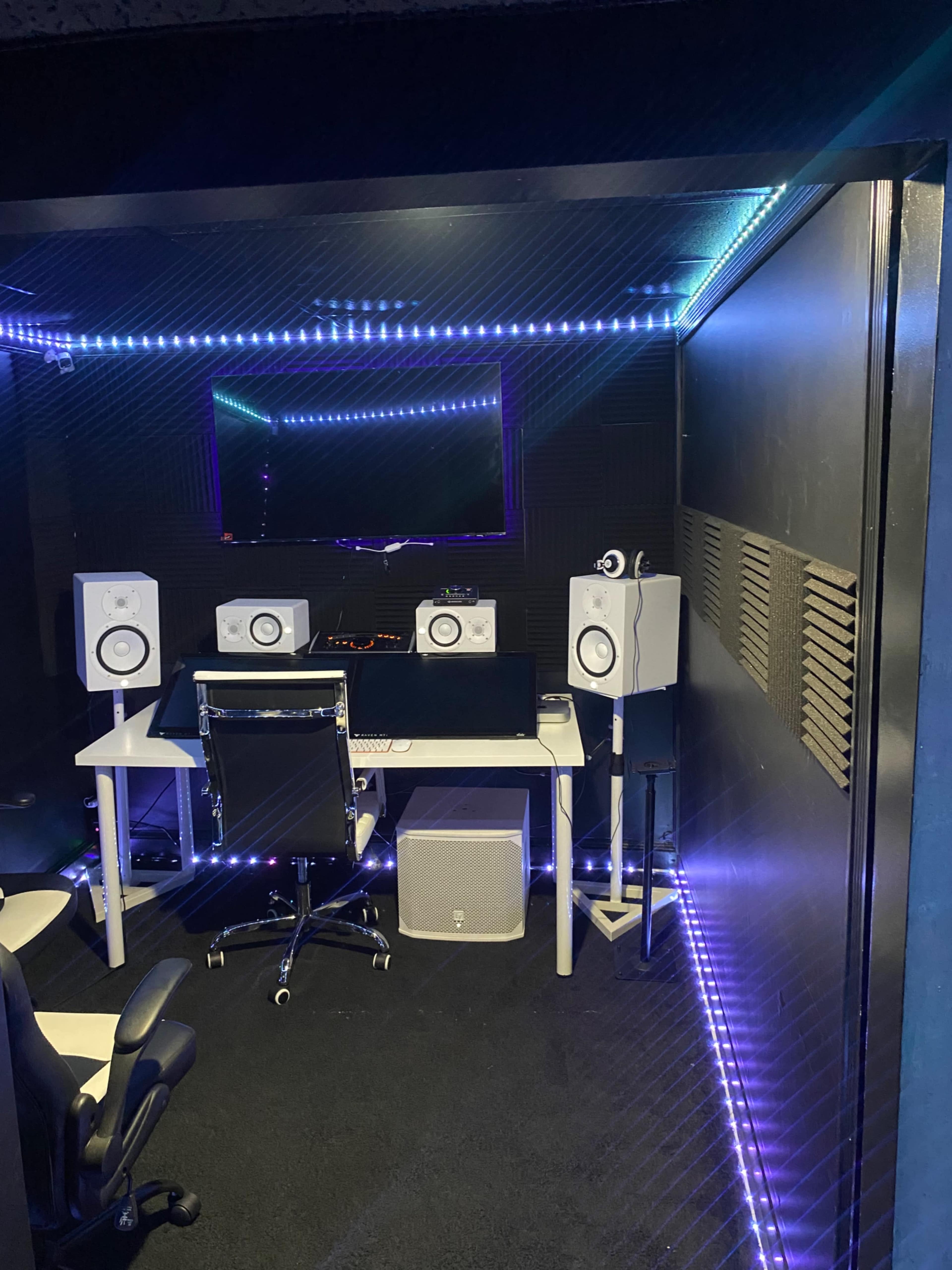A modern music studio with a desk, monitors, speakers, and acoustic treatment along the walls, illuminated by LED lights.