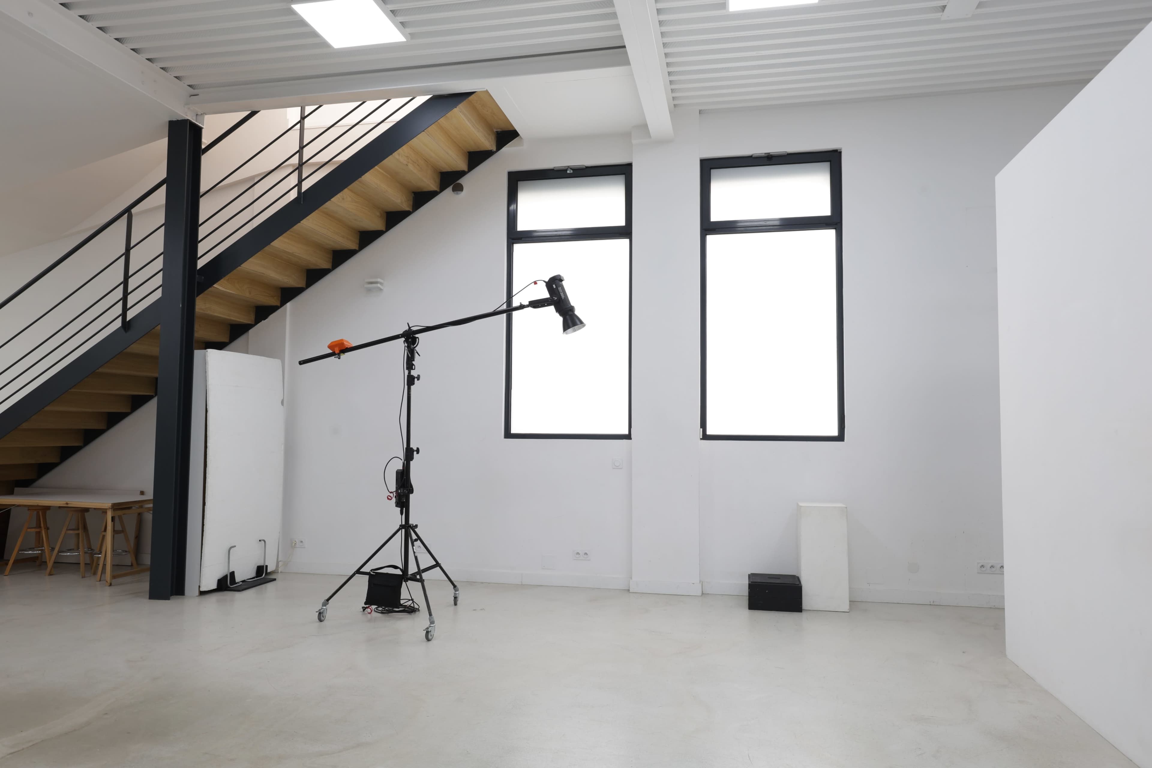 The image shows a spacious, well-lit room featuring a staircase, large windows, and a lighting stand on wheels positioned in the center.