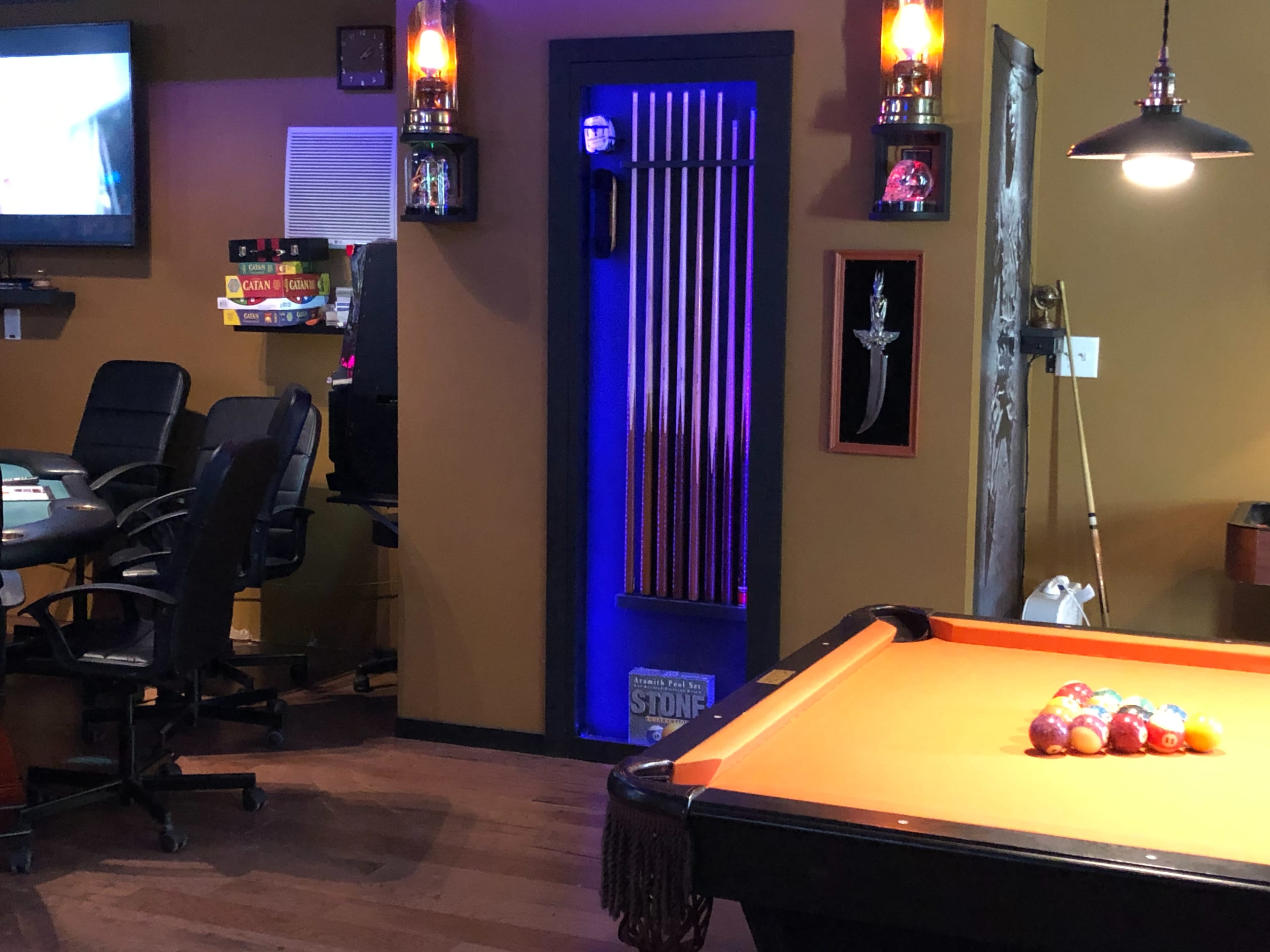 A game room with a pool table in the foreground, a poker table in the background, and a doorway lit with blue light.