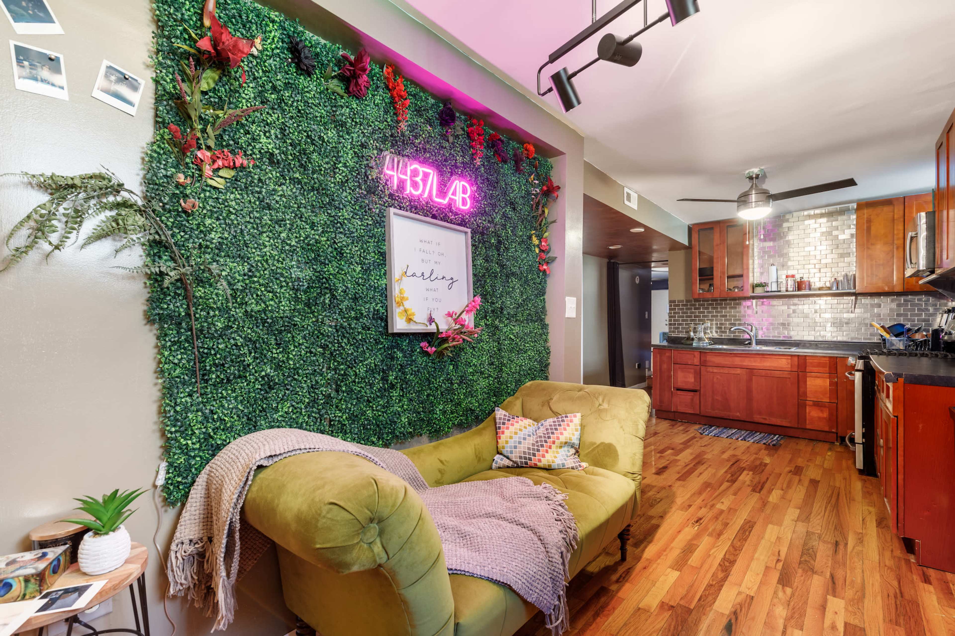 A cozy living space featuring a green wall of faux foliage with a neon sign, a yellow couch with a throw blanket and pillows, and a kitchen area in the background with wooden cabinetry.