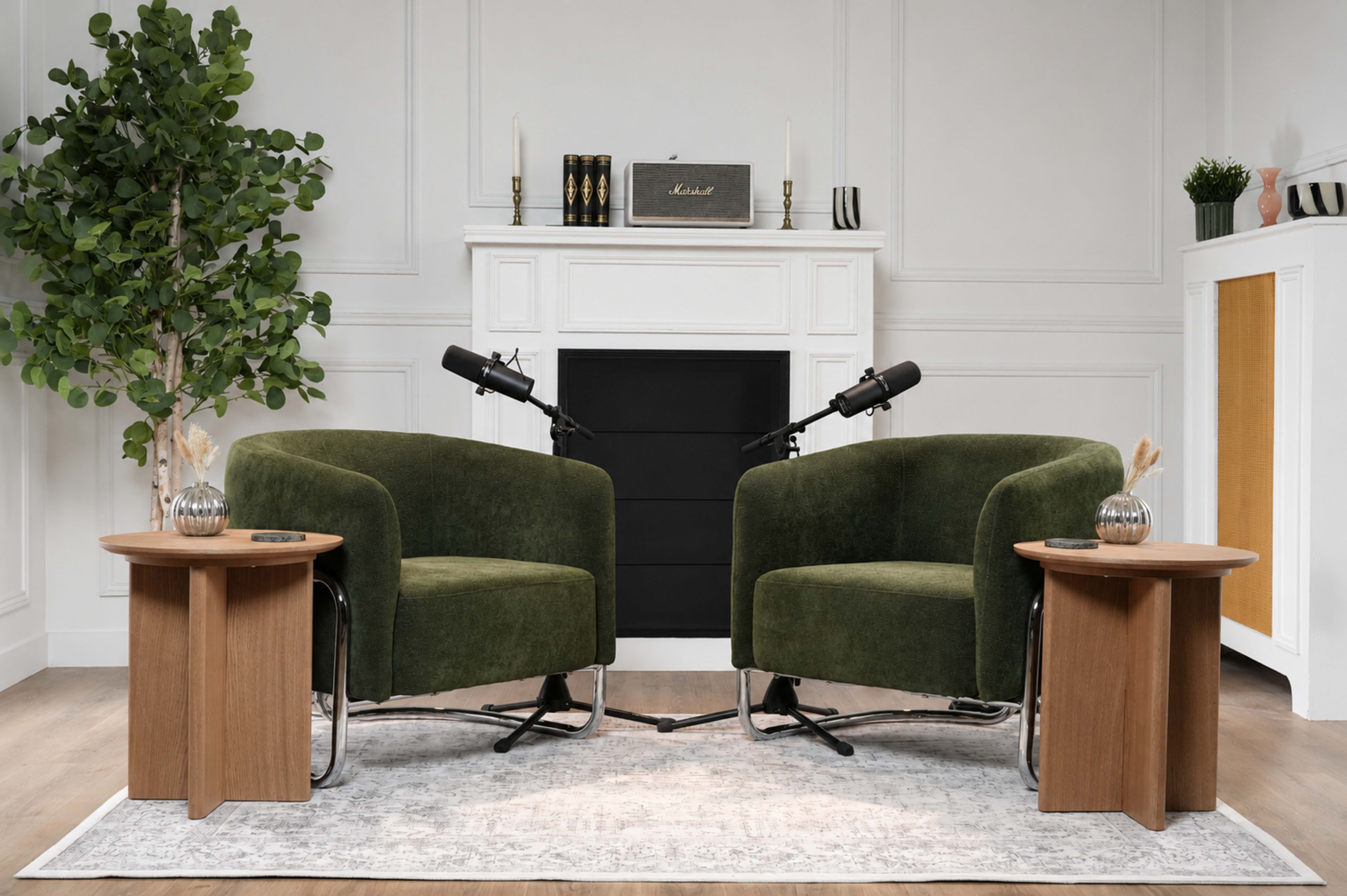 The scene features two green chairs placed opposite each other in front of a fireplace, with wooden side tables on either side and microphones set up between the chairs.