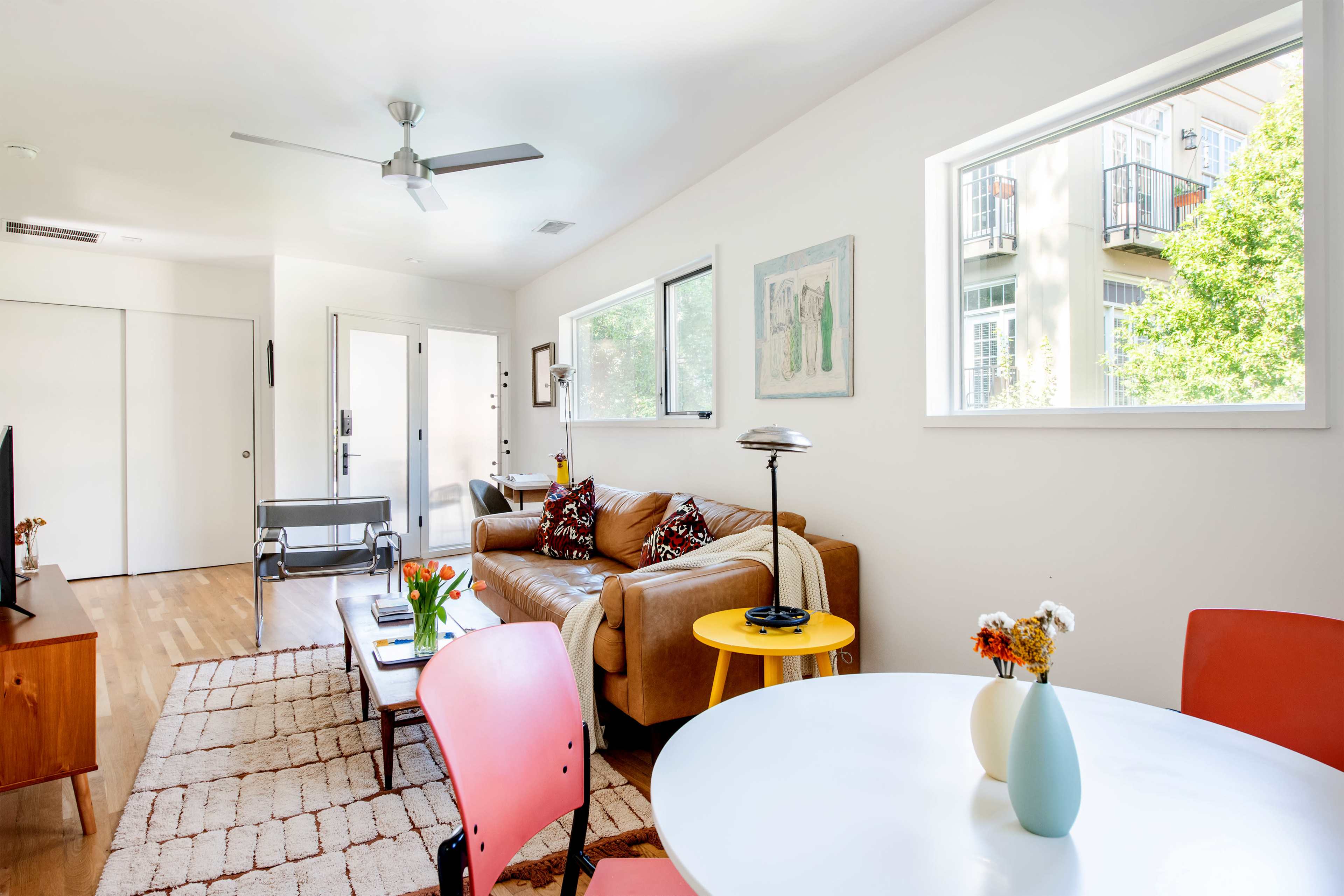 The image showcases a modern living room with a brown sofa, a round white dining table, and large windows that provide natural light.