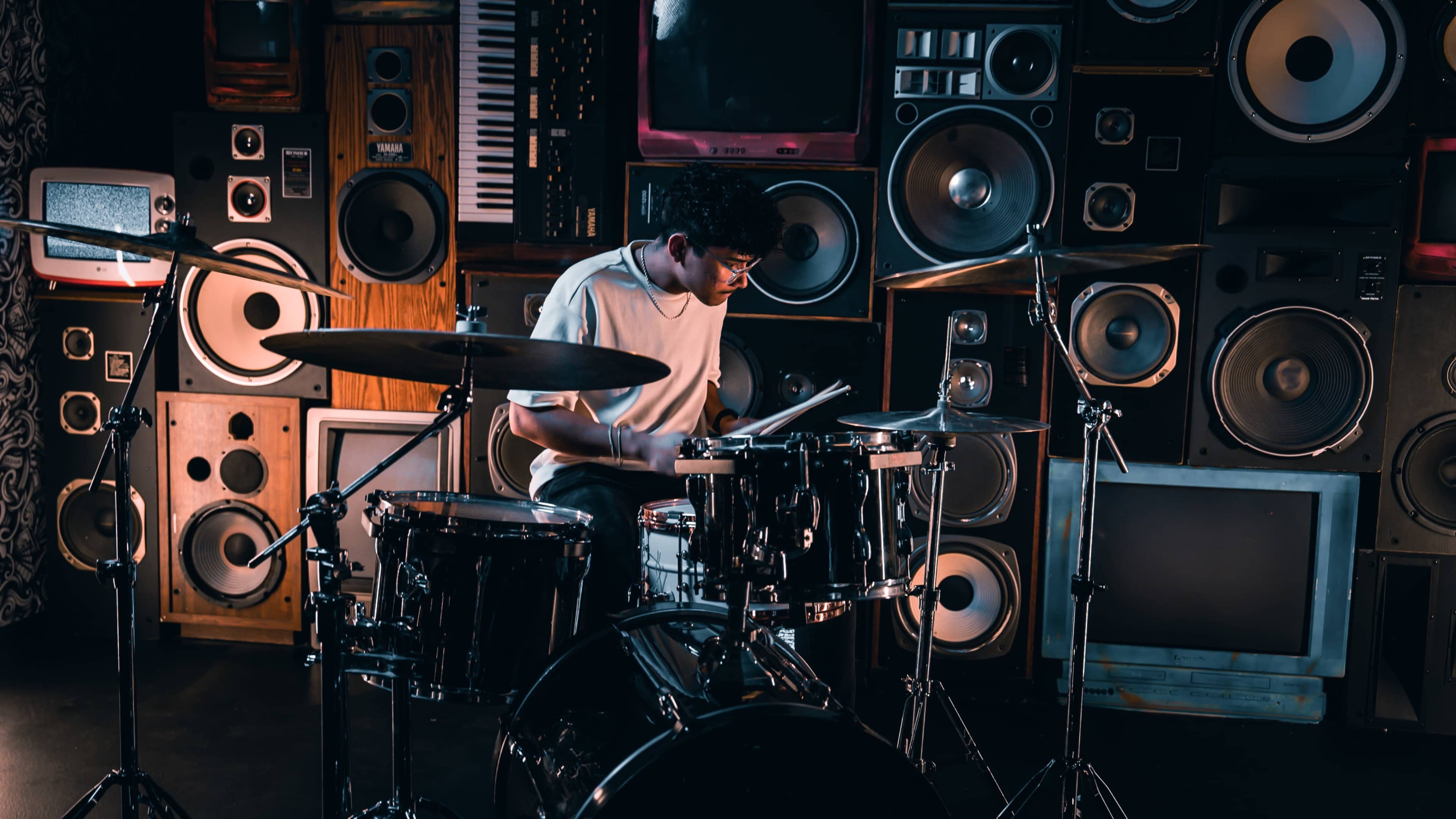 A drummer plays a set of drums surrounded by an array of vintage speakers and musical instruments.