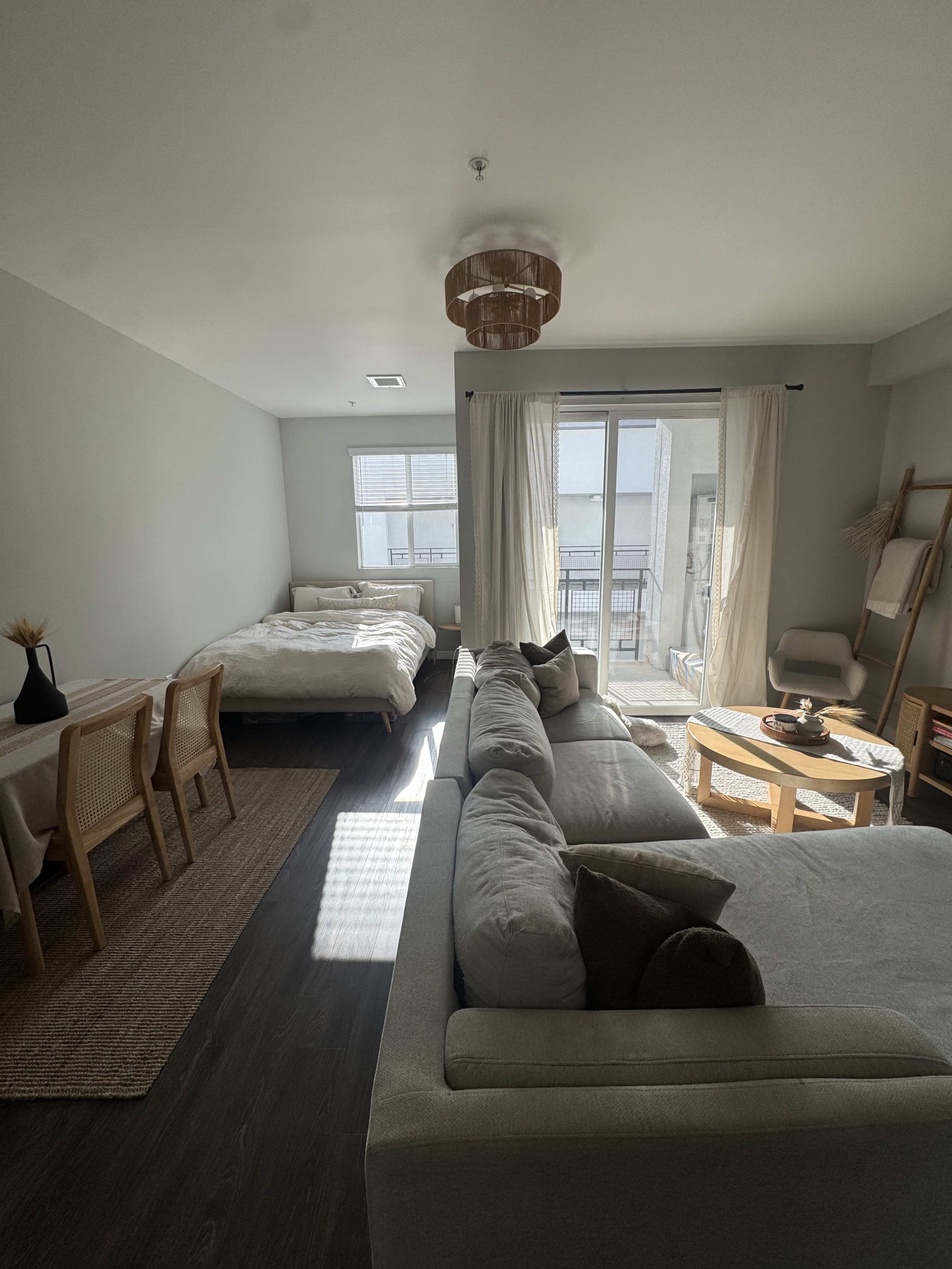 The image shows a small living space with a sofa, a dining area, a bed, and a balcony door, all featuring neutral-colored decor.