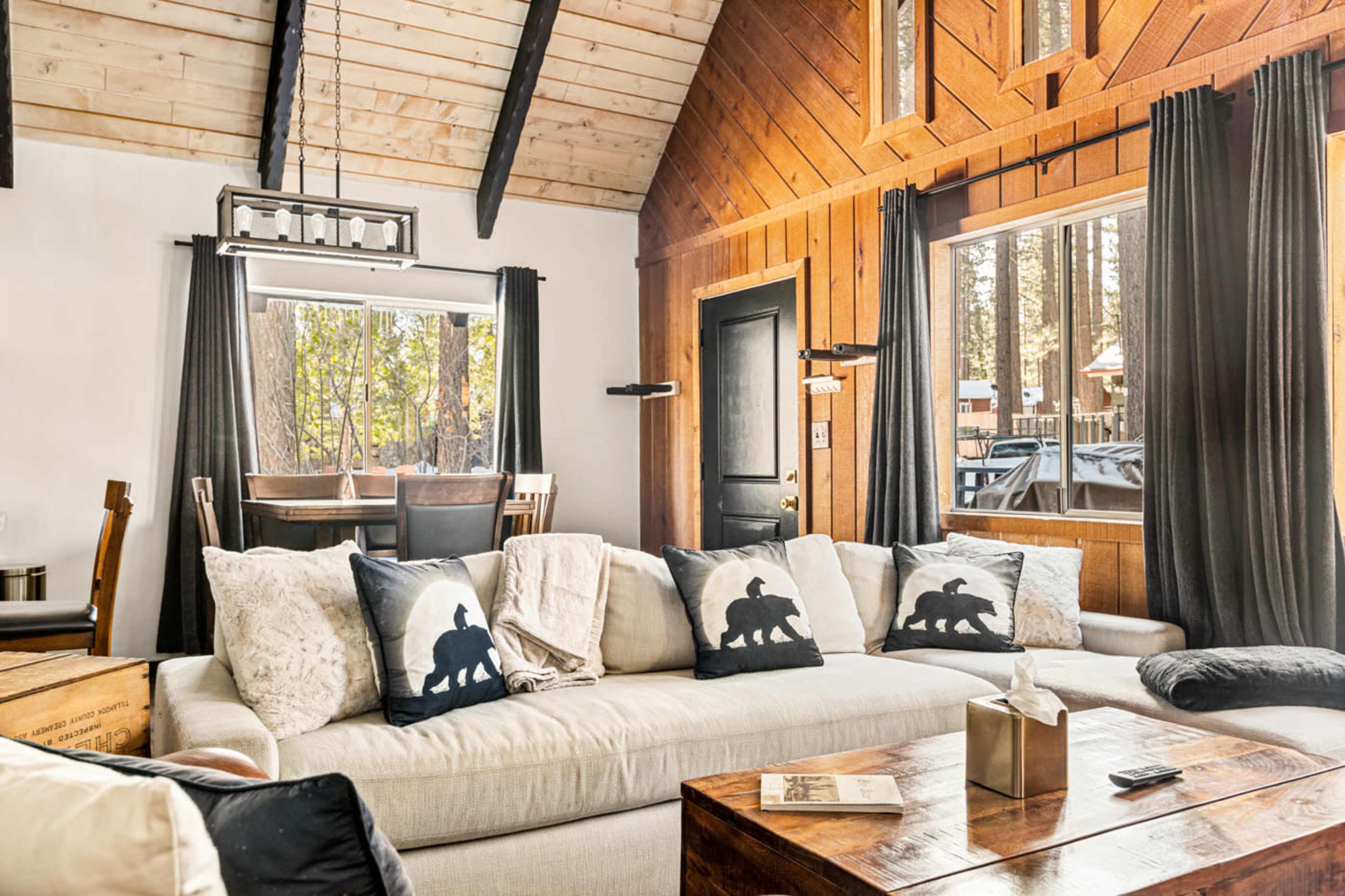 A cozy living room features a white sofa adorned with bear-themed throw pillows, wooden accents, and large windows letting in natural light.
