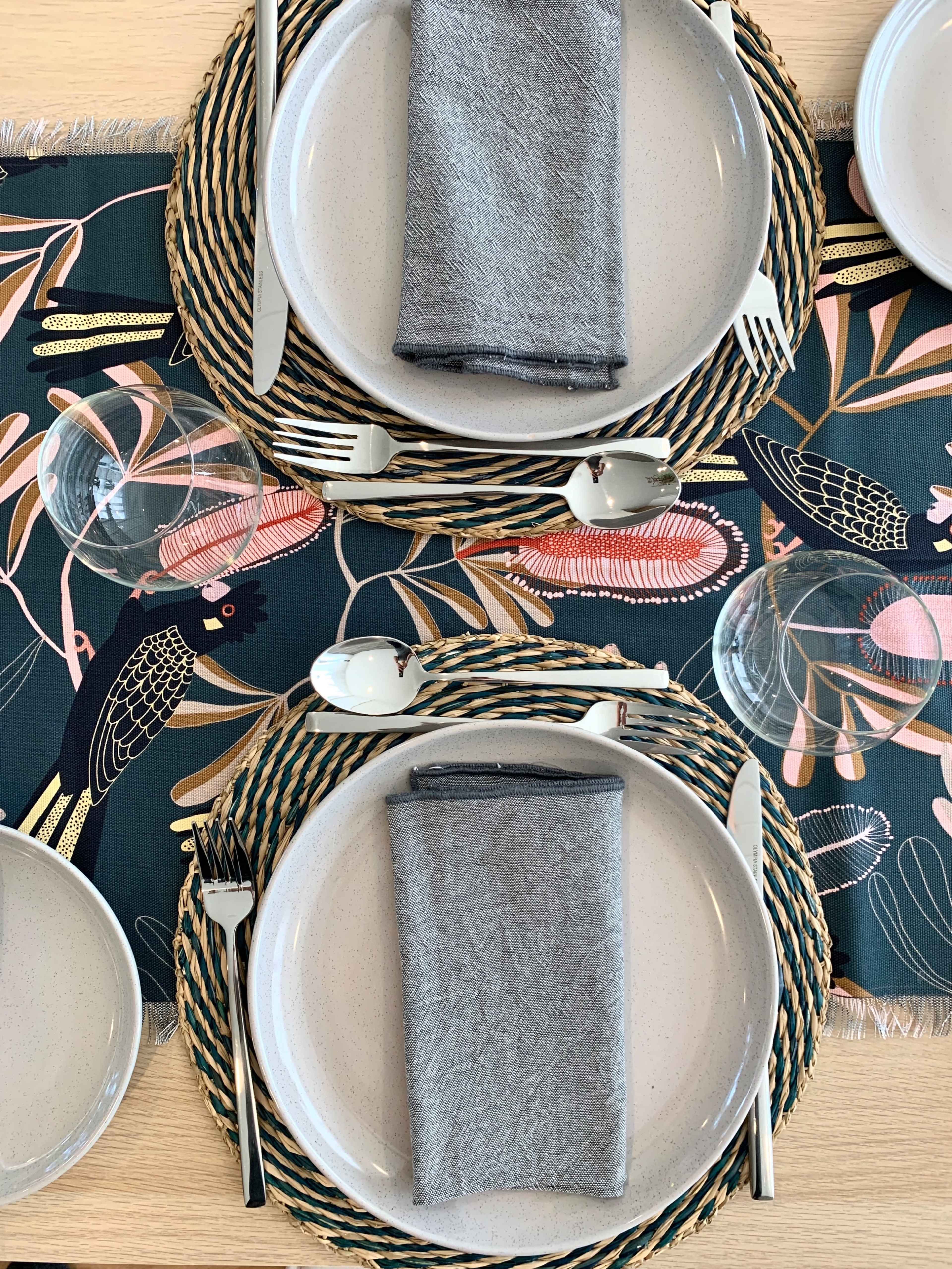 A dining table set with two plates, gray napkins, cutlery, and glasses, arranged on a patterned table runner.