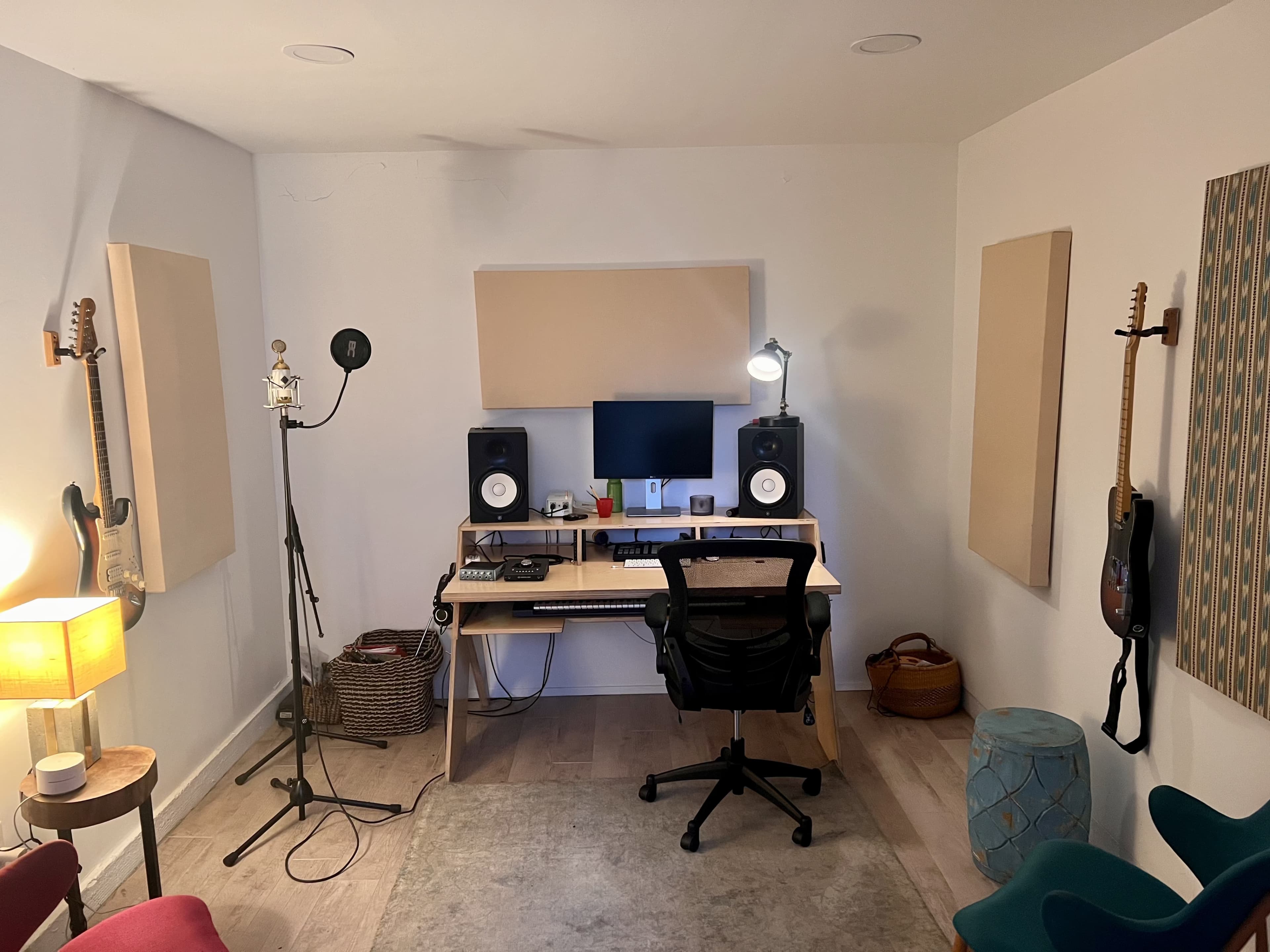 The image shows a small music studio with a desk, computer, speakers, a microphone on a stand, and guitars mounted on the walls.
