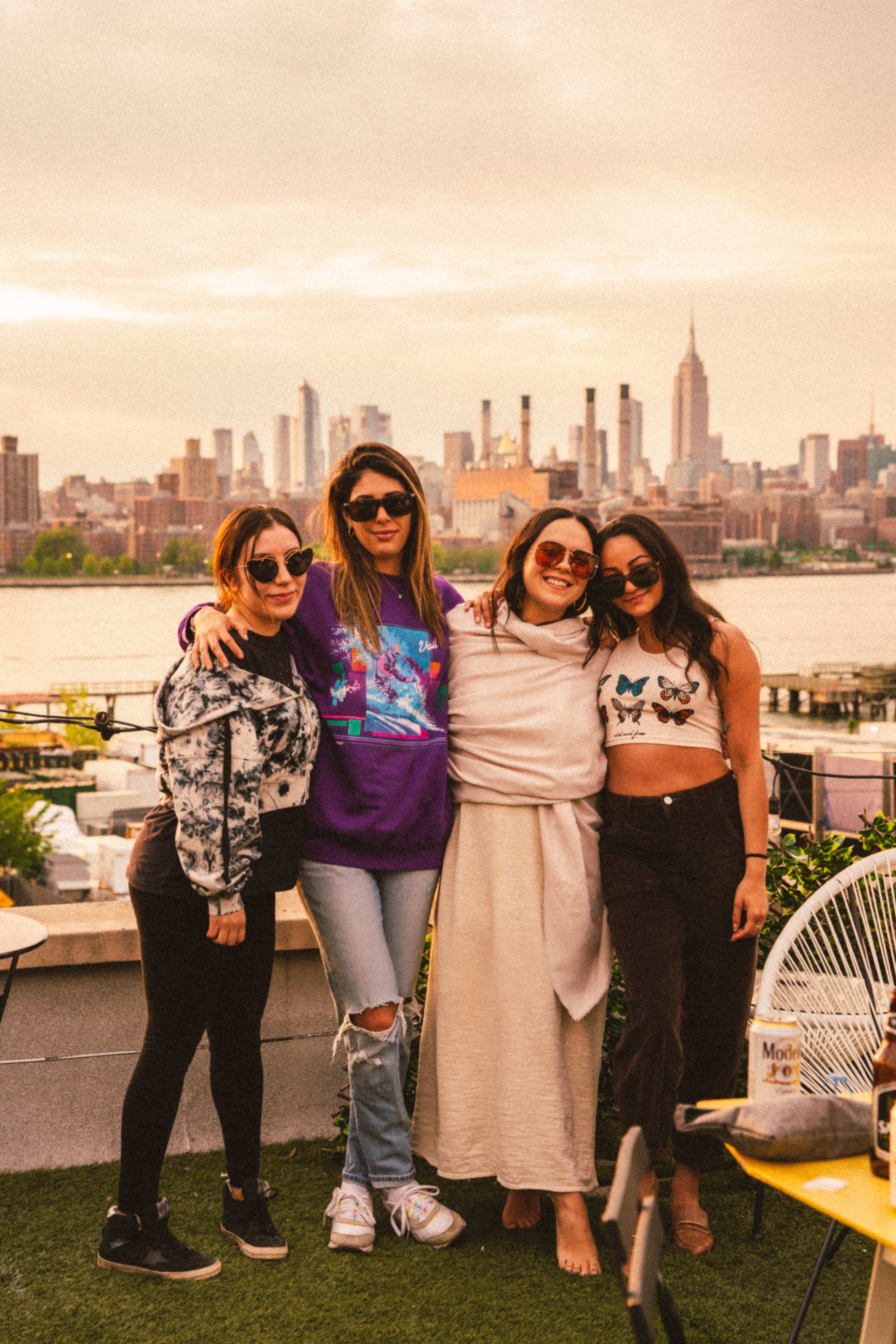 Four women pose together on a rooftop with the New York City skyline in the background at sunset.