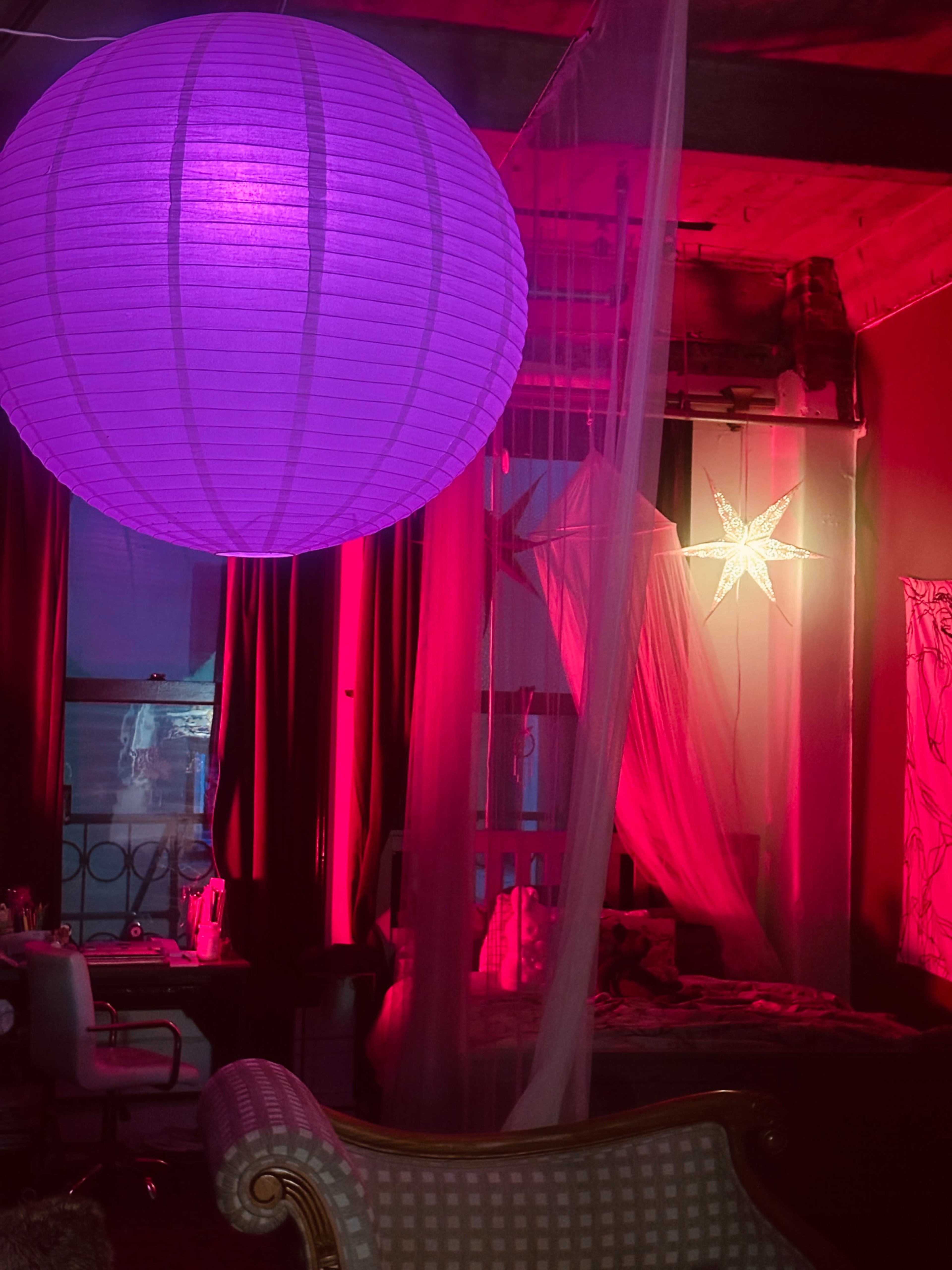 A large purple paper lantern illuminates a room with red and pink lights, featuring a canopy bed and a decorative star hanging from the ceiling.