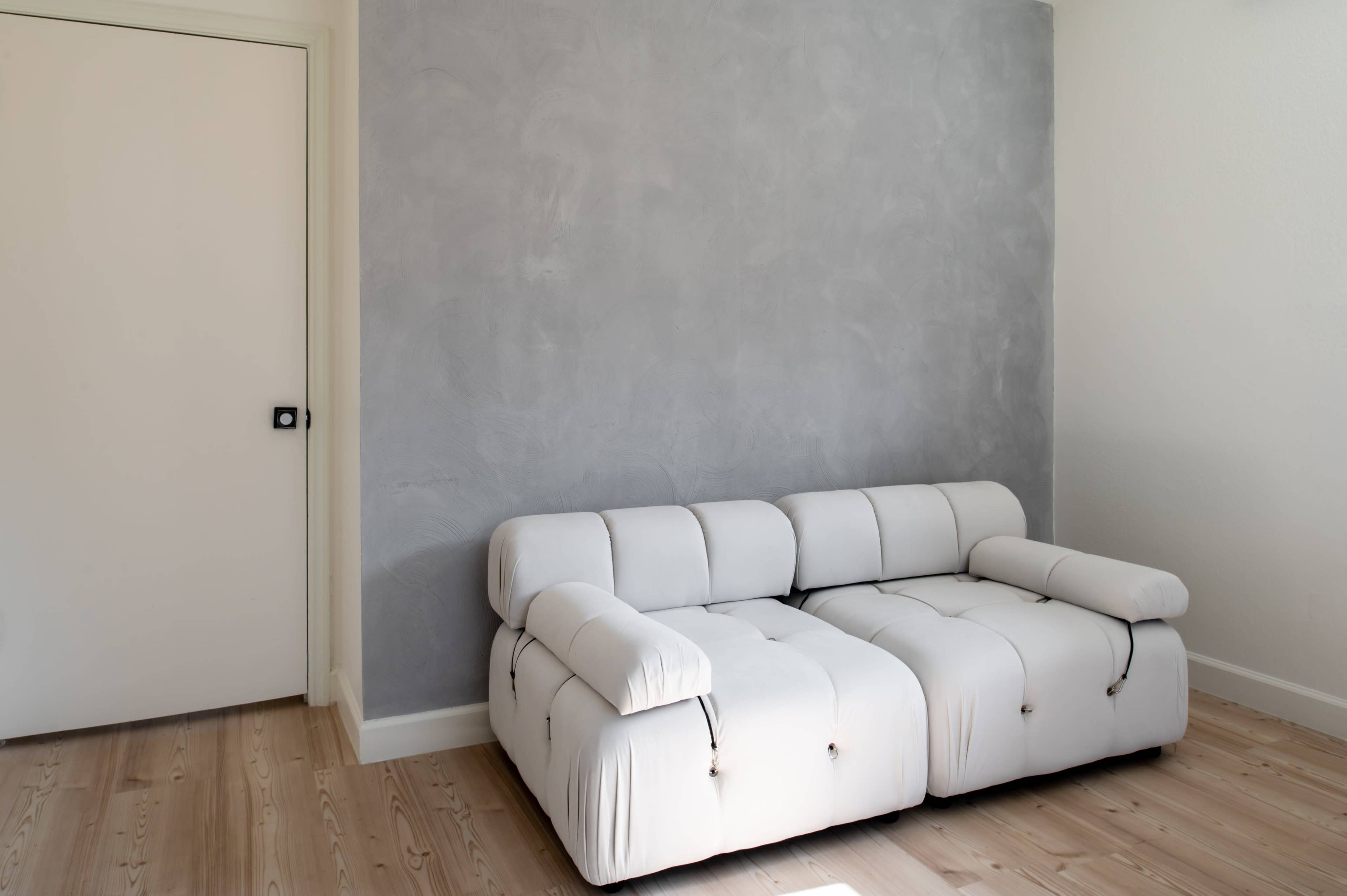 A white, tufted sofa is positioned against a gray wall in a minimalistic room with light wood flooring.