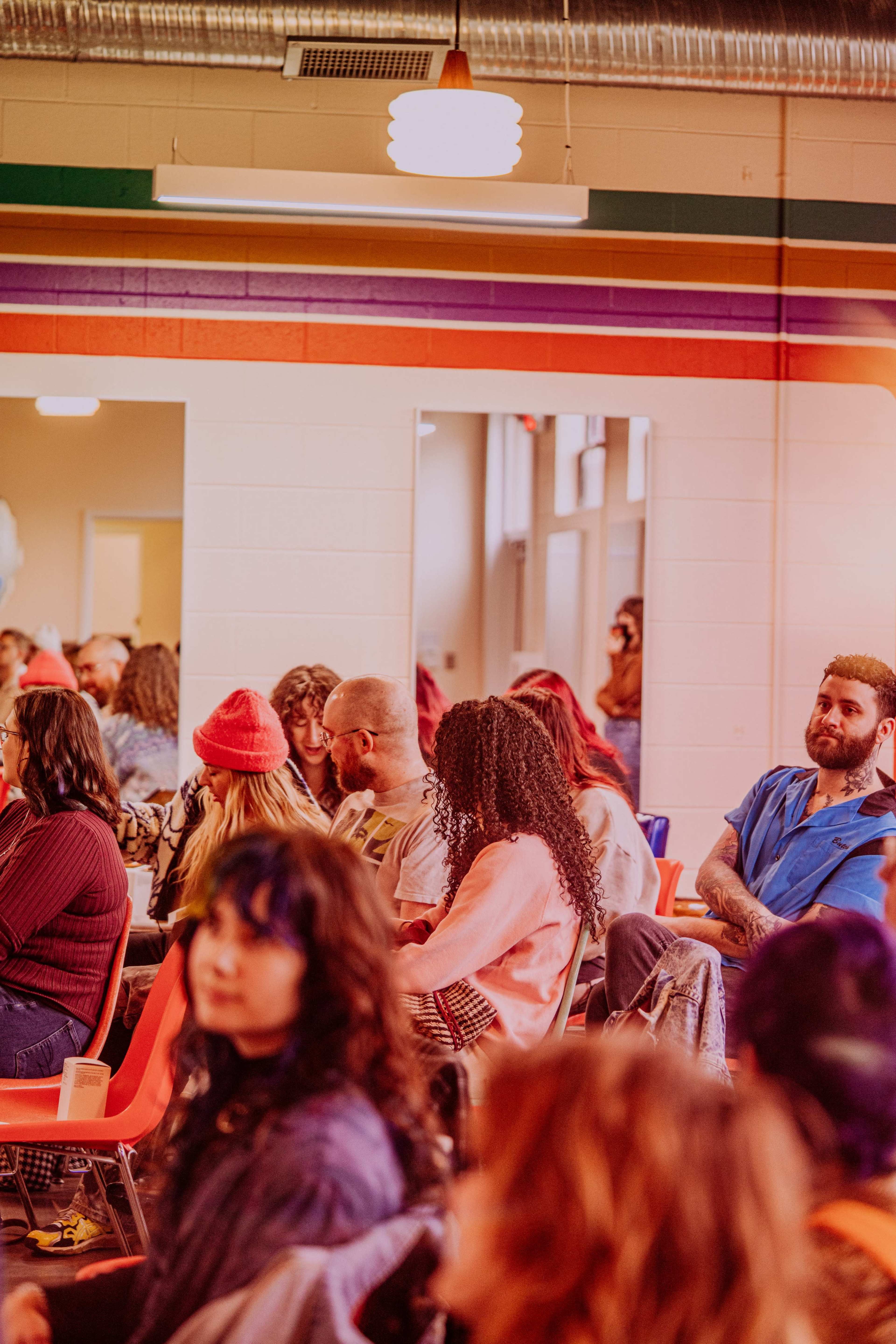A diverse group of people sits in a brightly lit room with colorful wall stripes, listening attentively.