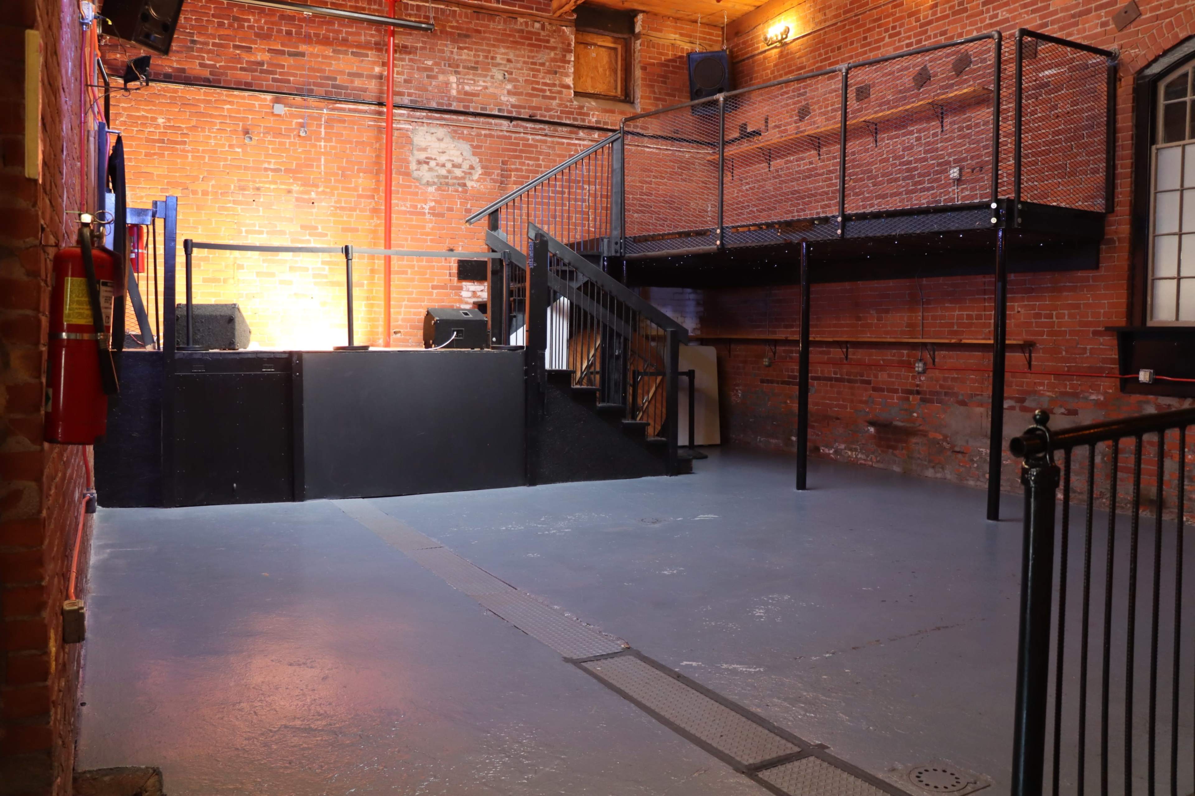 The image shows an empty venue with exposed brick walls, a raised platform, and a staircase, illuminated by warm lighting.