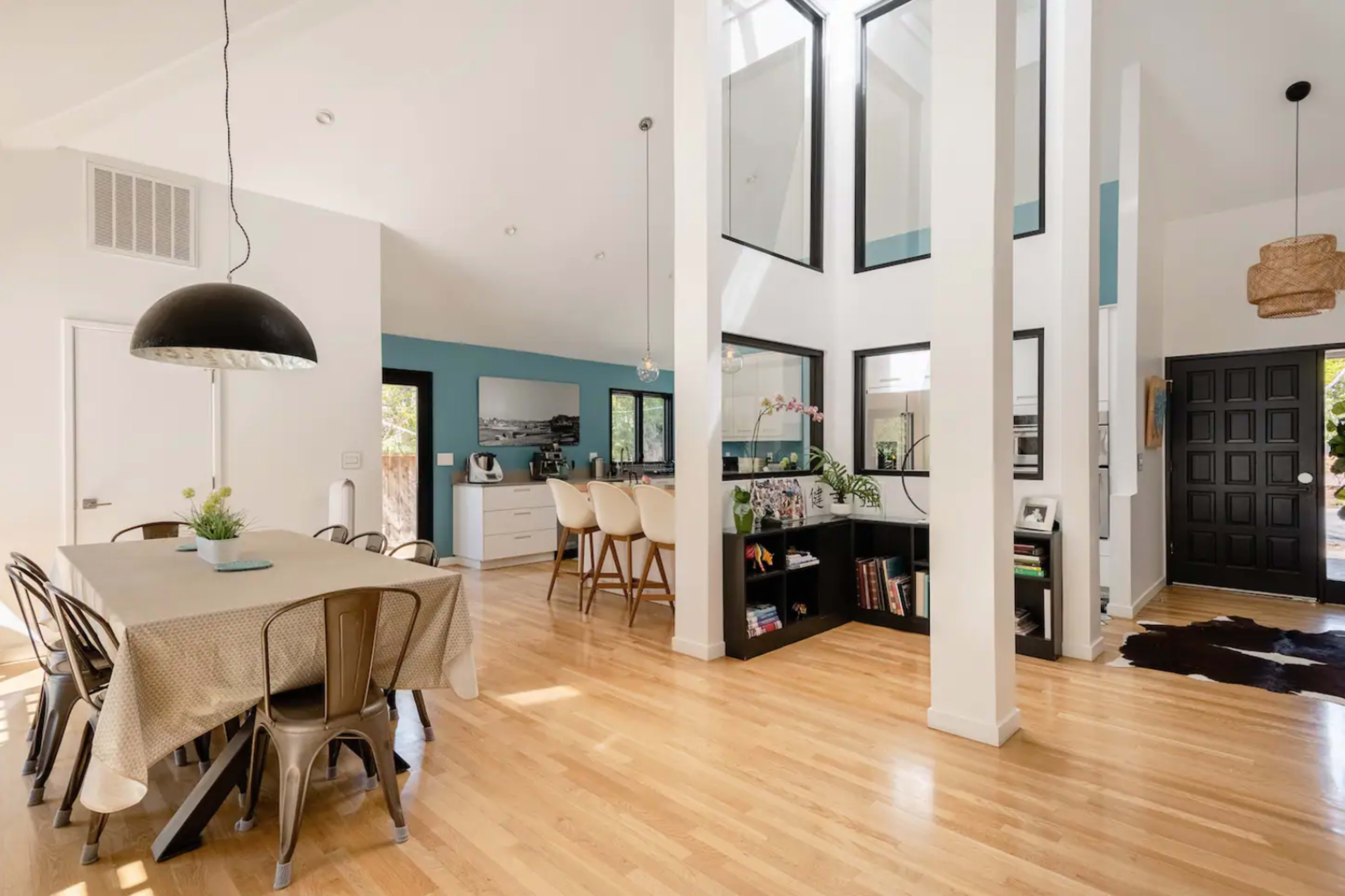 A modern open-concept living space with a dining area, kitchen, and a black door, characterized by large windows and hardwood flooring.