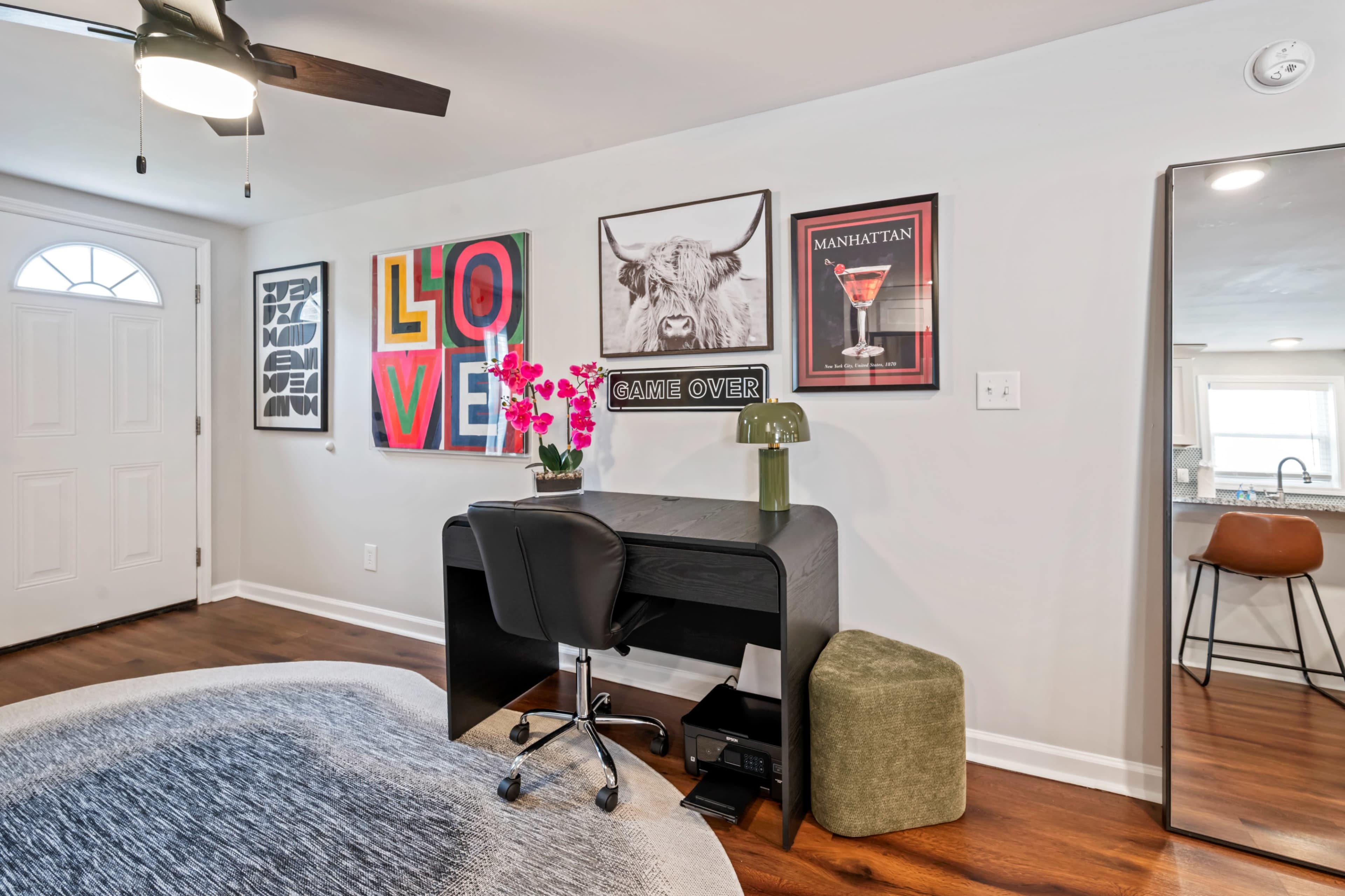 The image shows a modern workspace featuring a black desk with a chair, surrounded by colorful framed art on the wall, a plant, and a mirror nearby.